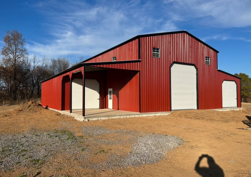 Red metal barn with large white doors on a dirt lot under a blue sky.