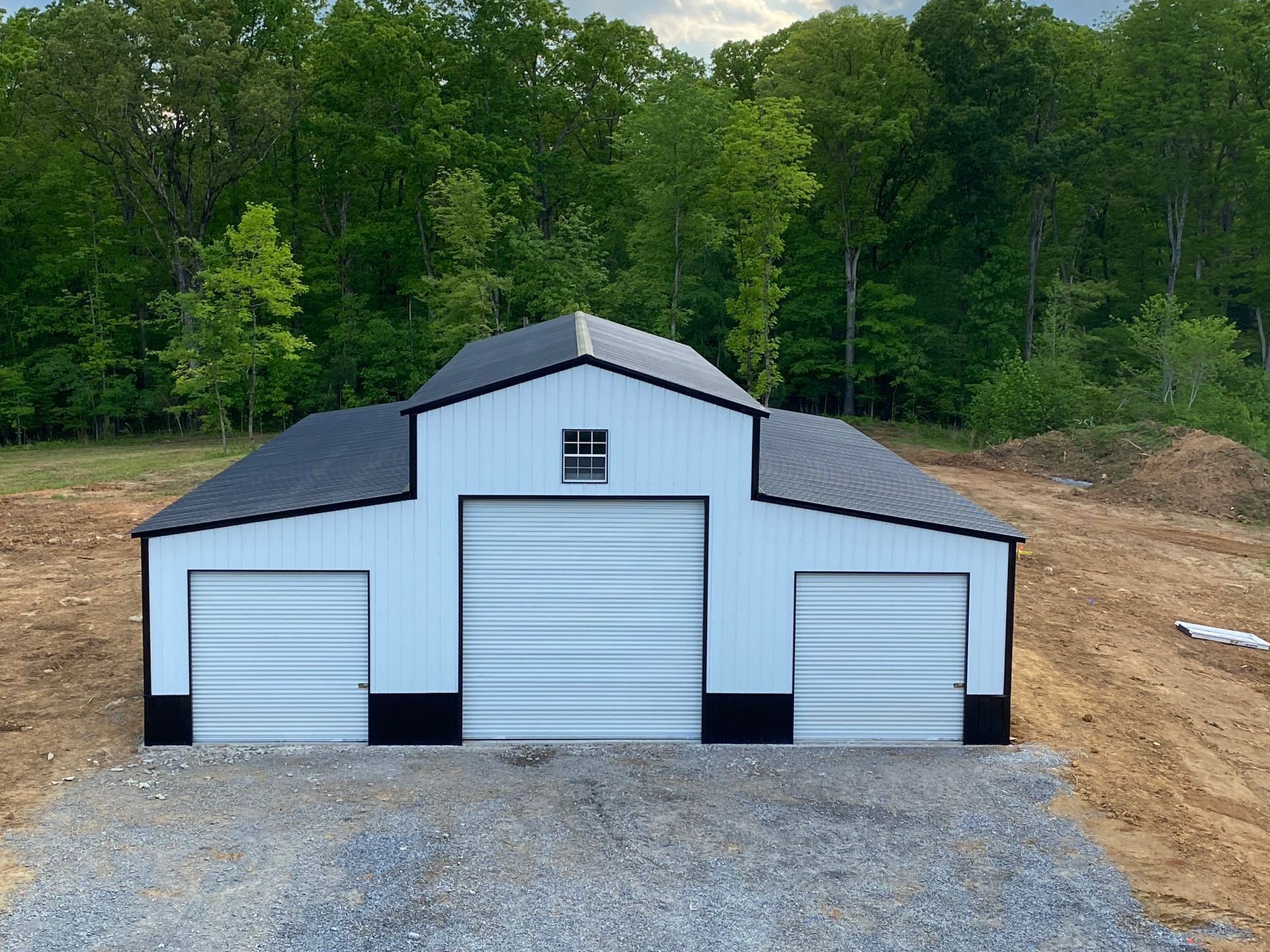 Three-door white garage with black roof on a gravel pad, backed by a wooded hillside.