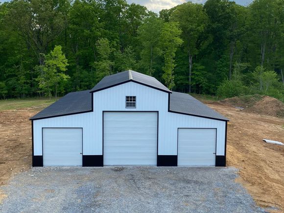 White metal garage with three roll-up doors on a gravel lot, backed by trees and a dirt slope