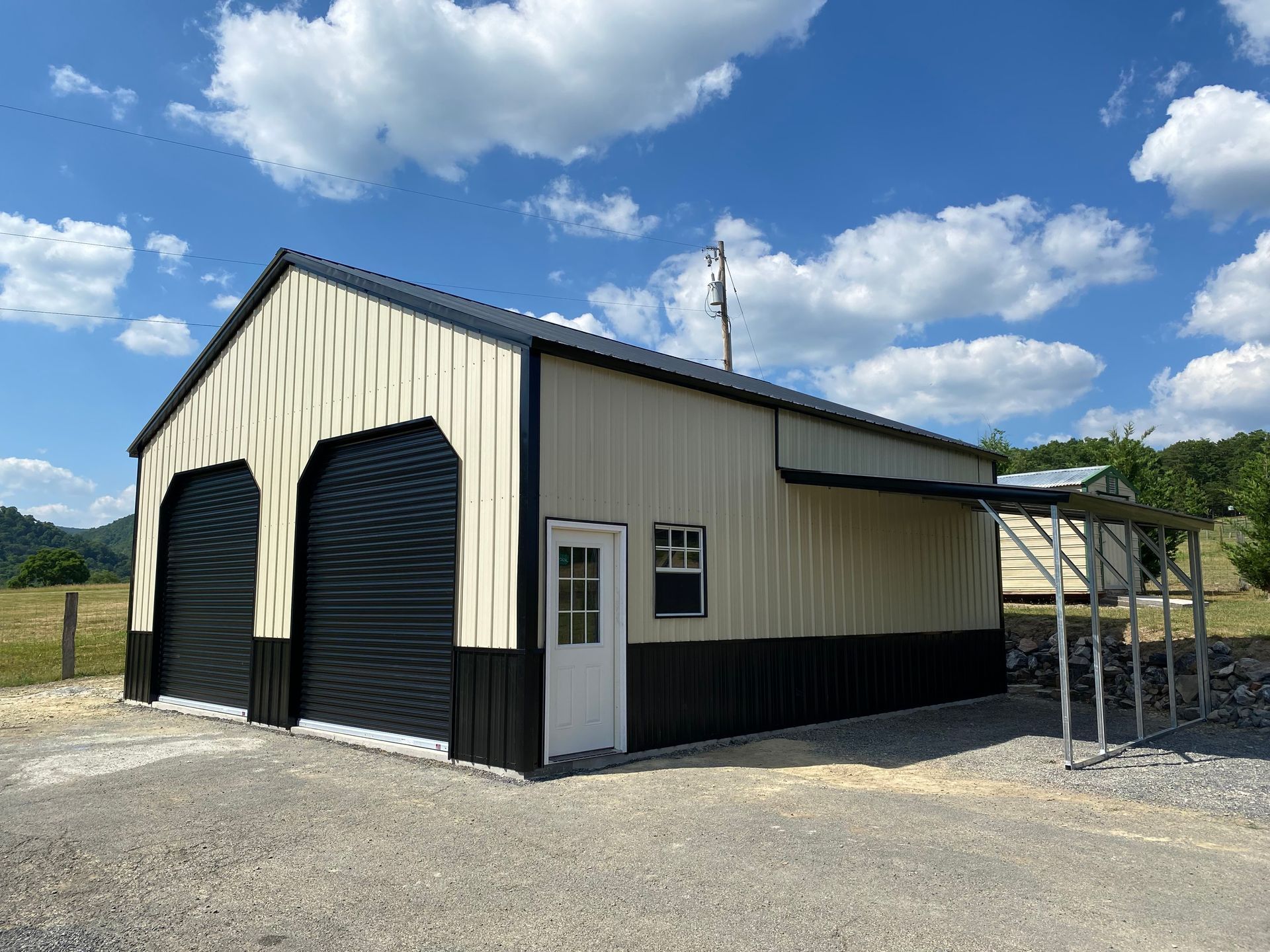 Small beige metal garage with black doors on a gravel lot under a blue sky with clouds