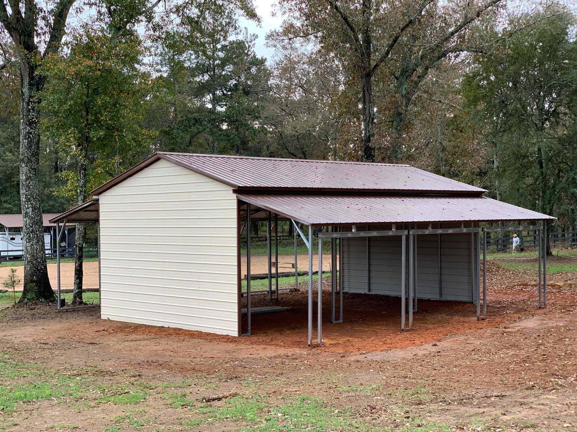 Small beige metal carport shed with open sides in a wooded yard with dirt ground