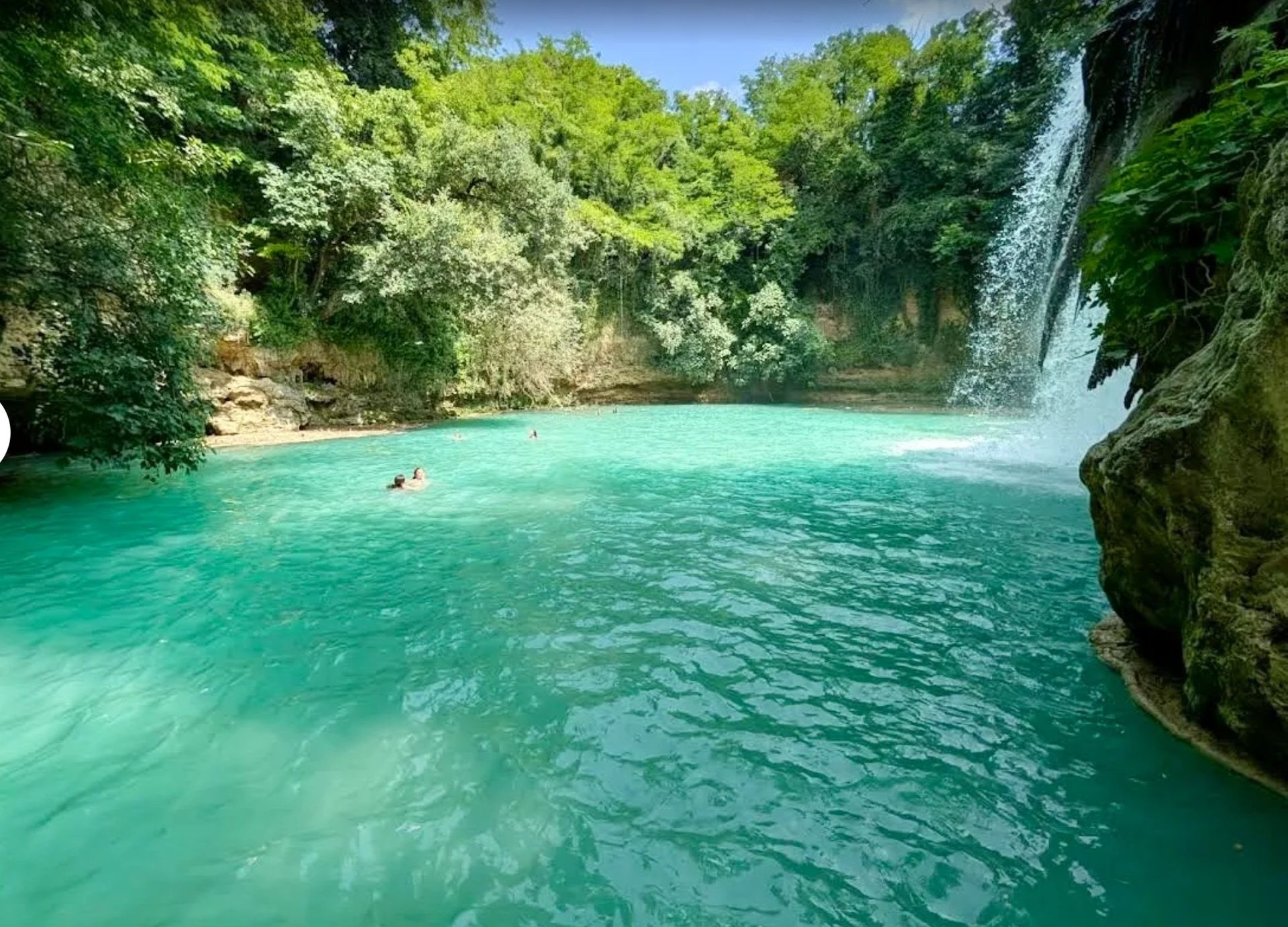 Turquoise tropical pool with a small waterfall, surrounded by lush green forest and rocks