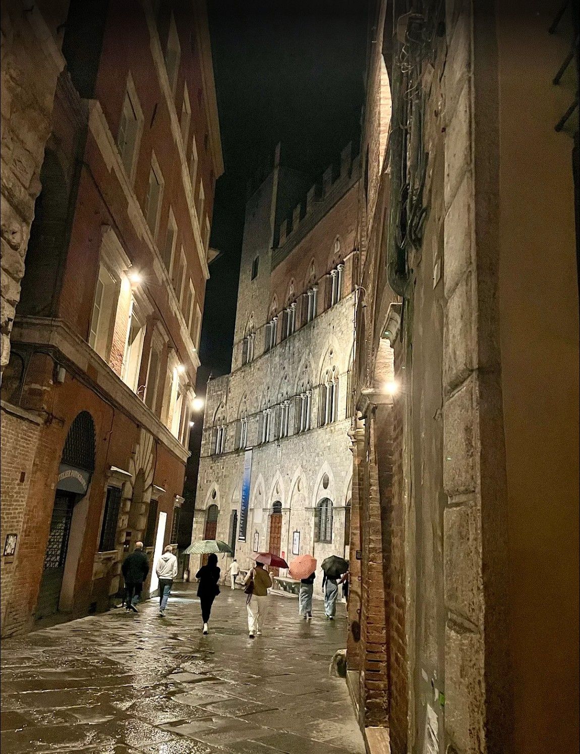 Narrow historic alley at night, with people walking on wet cobblestones between tall old buildings.