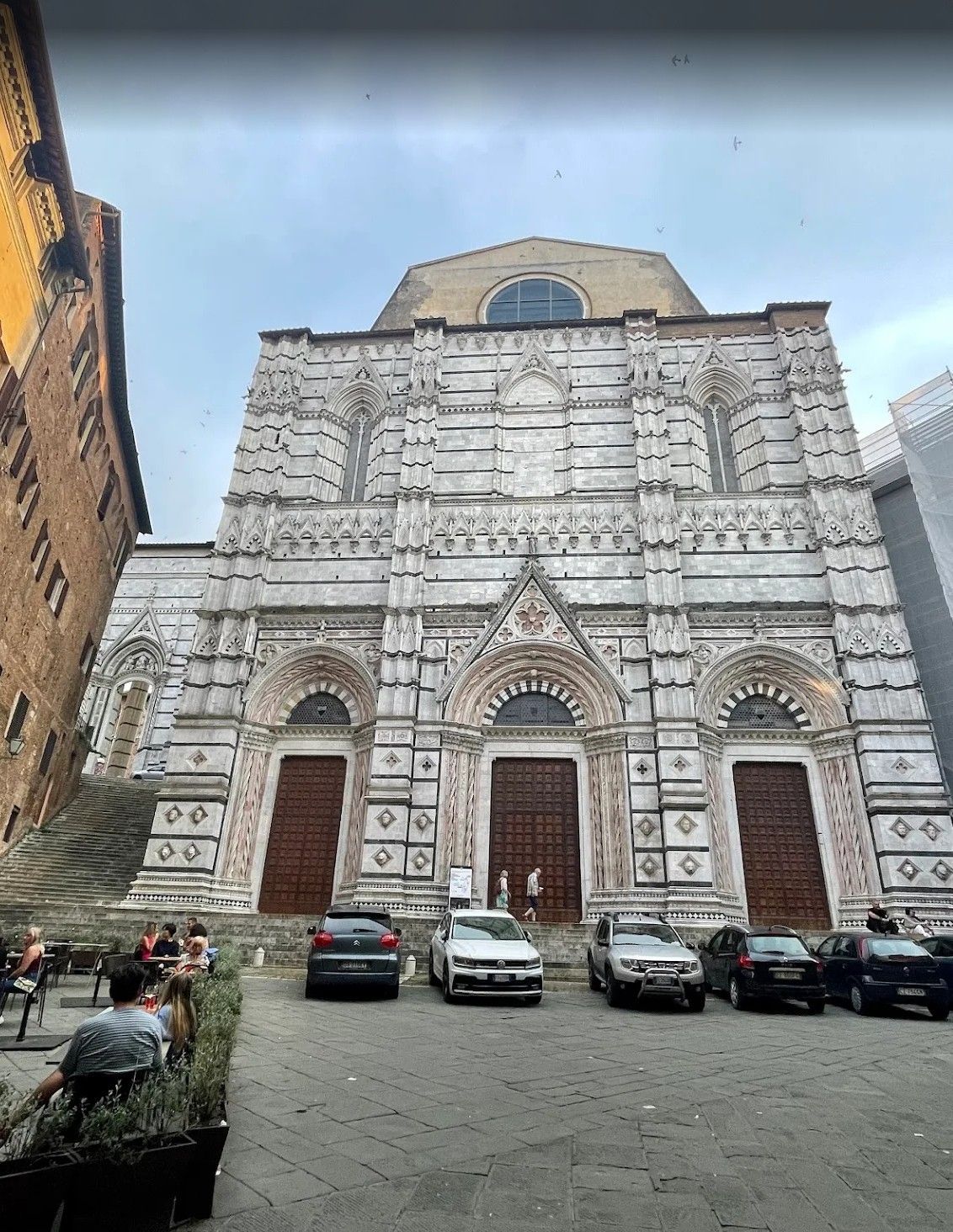 Ornate white stone cathedral facade with three arched doors and parked cars in a plaza