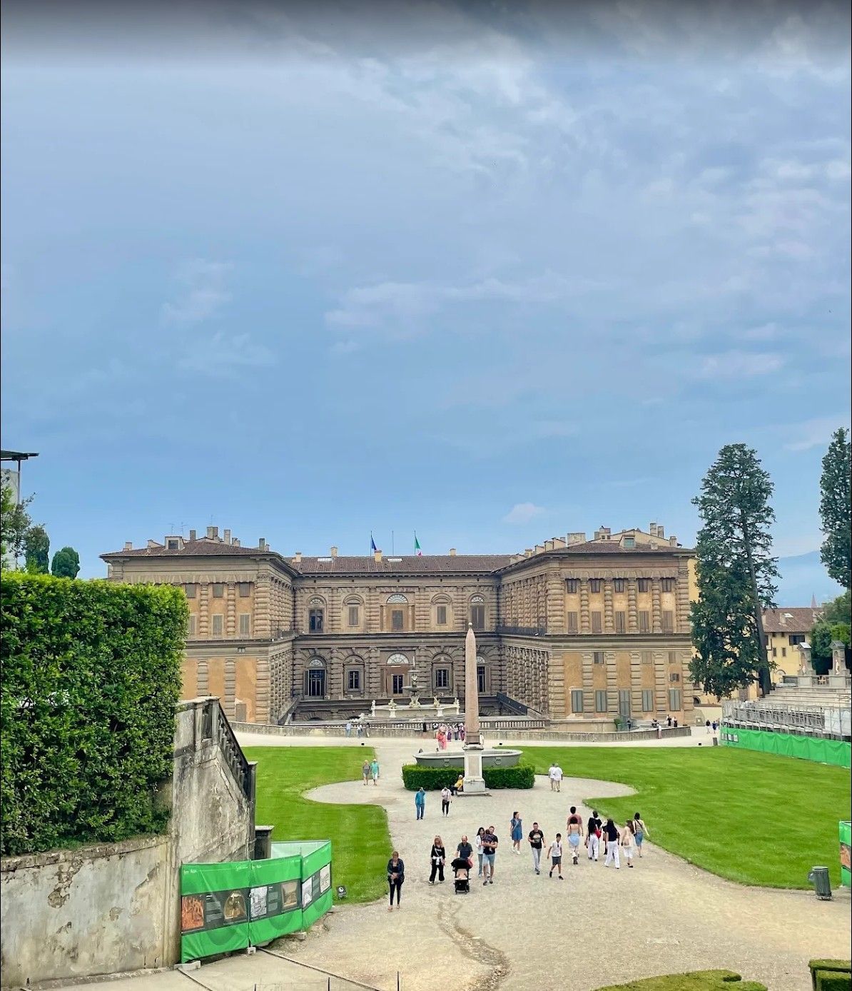 Historic stone palace with a central courtyard, green lawns, and visitors under a cloudy sky.