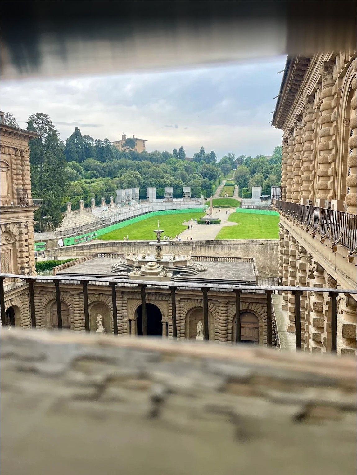 Stone palace courtyard with green lawns and a fountain, viewed through an ornate terrace opening
