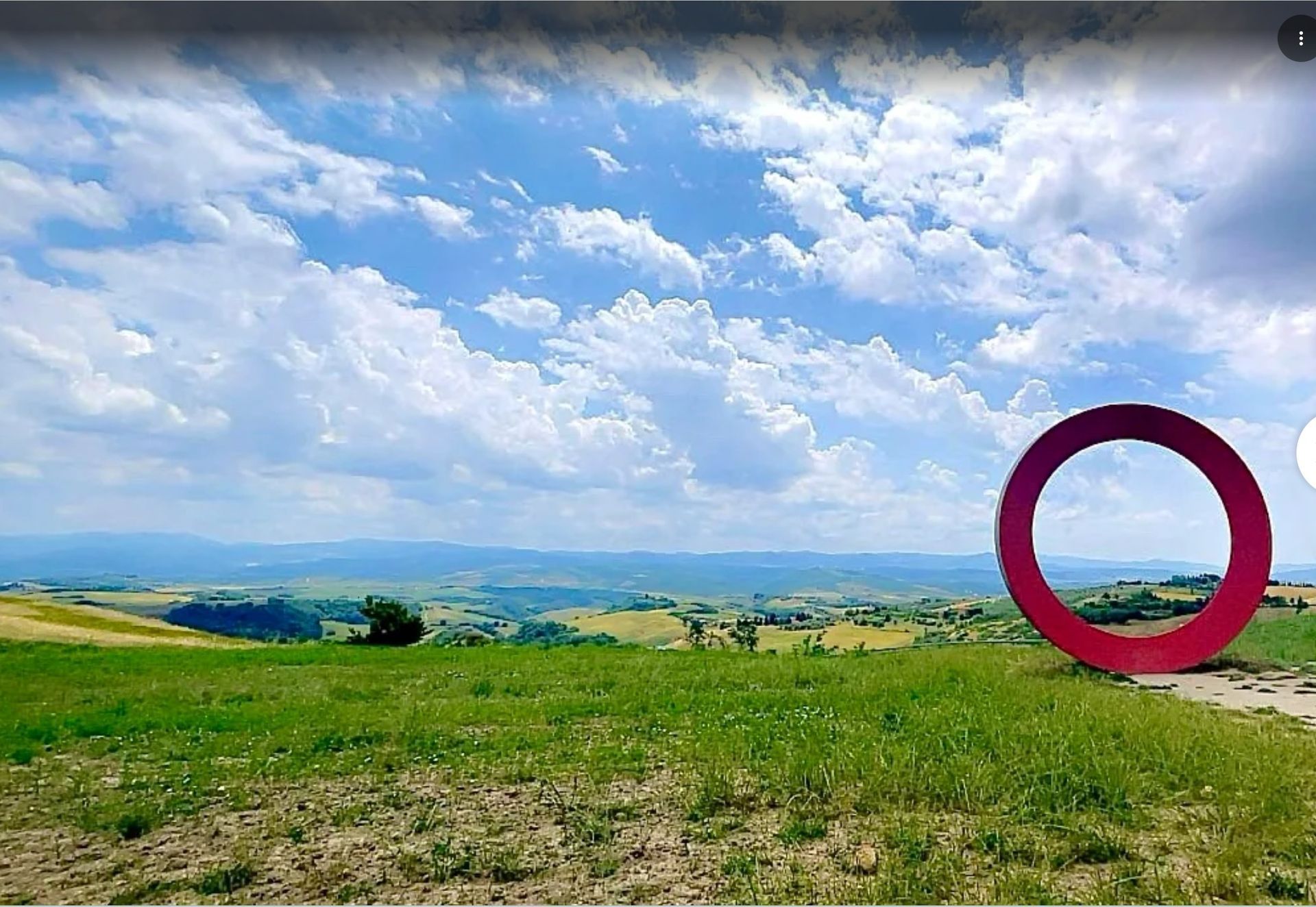 Grassy hilltop overlooking a wide valley under a cloudy blue sky, with a red circle marking the right side.