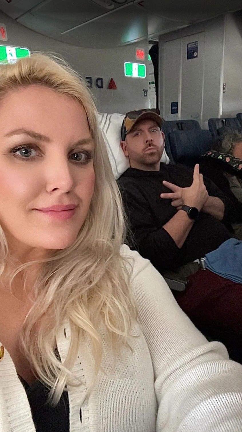 Two people seated on an airplane, one taking a selfie in the foreground.