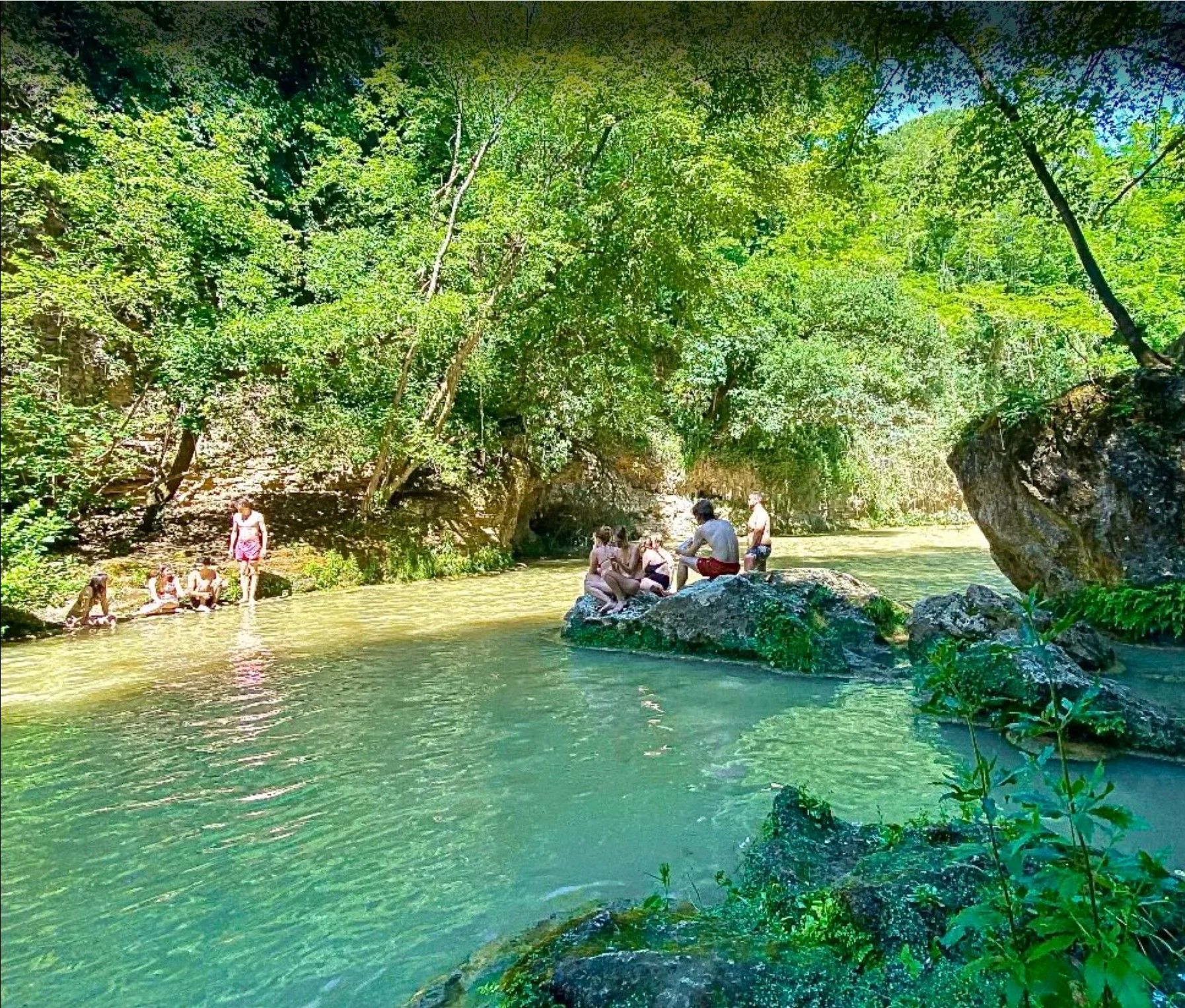People sitting on rocks by a turquoise forest pool, with one person wading nearby under lush green trees.