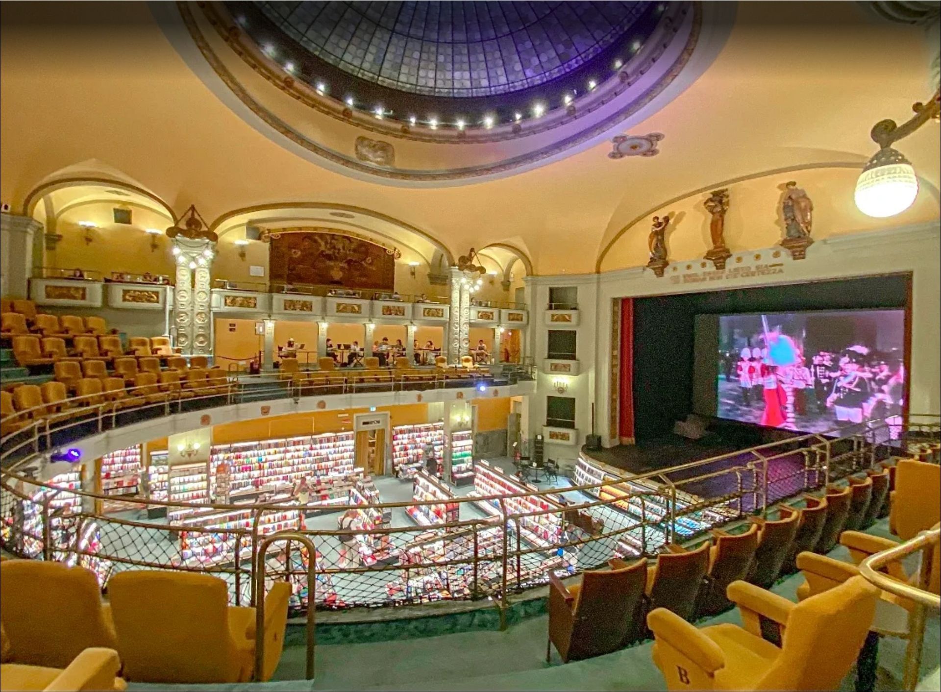 Empty theater interior with ornate balconies, yellow seats, and a large stage screen displaying pink visuals