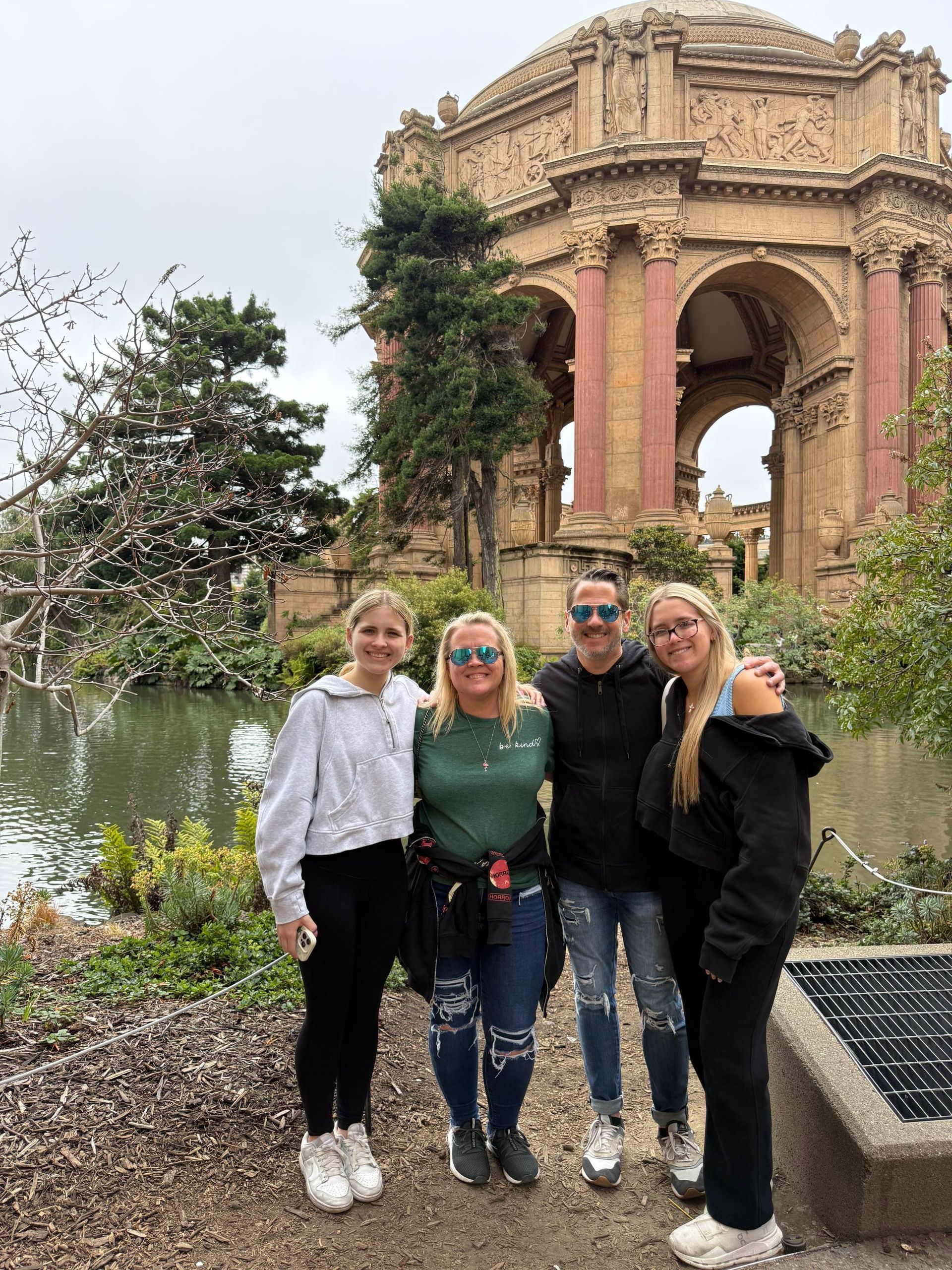Four people posing outdoors by a pink ornate arch beside a reflective pond and flowering trees.