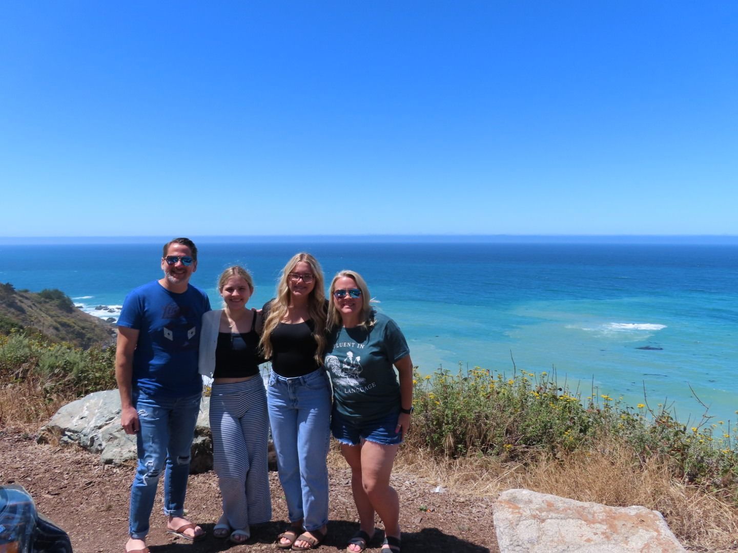 Four friends posing on a coastal cliff above a blue ocean under a clear sky