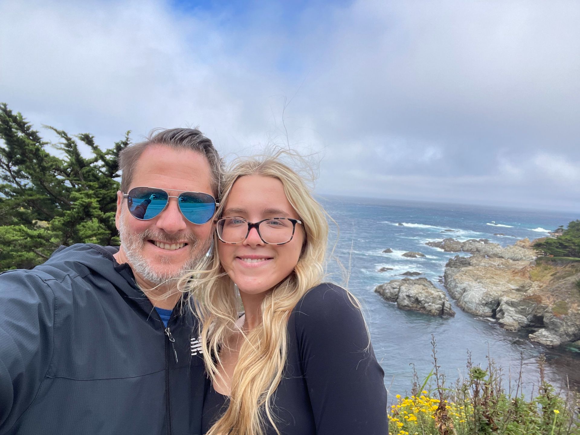 Two people smiling by a rocky ocean coast under a cloudy sky