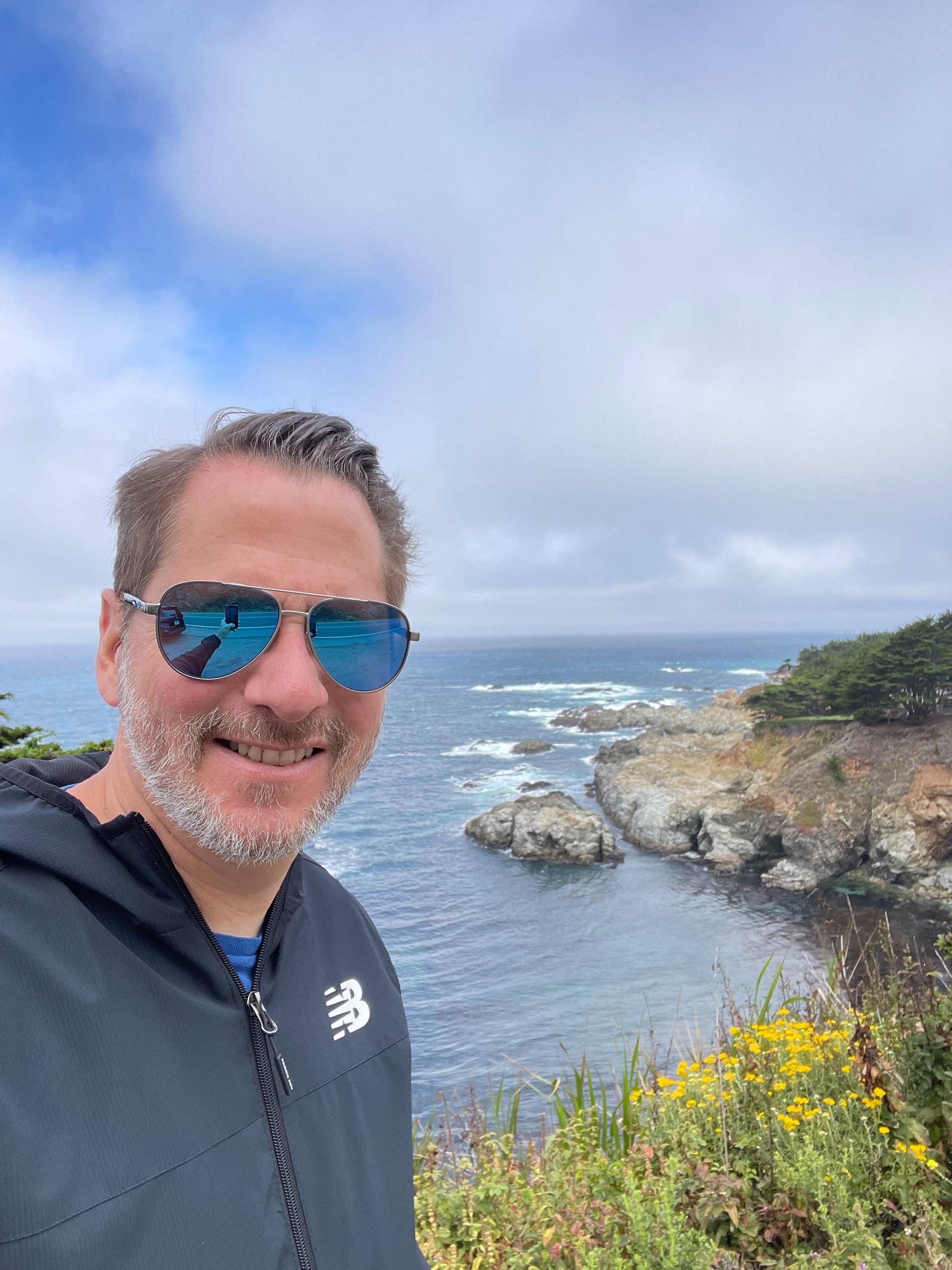 Person in blue sunglasses smiles at a rocky seaside cliff under a cloudy sky.