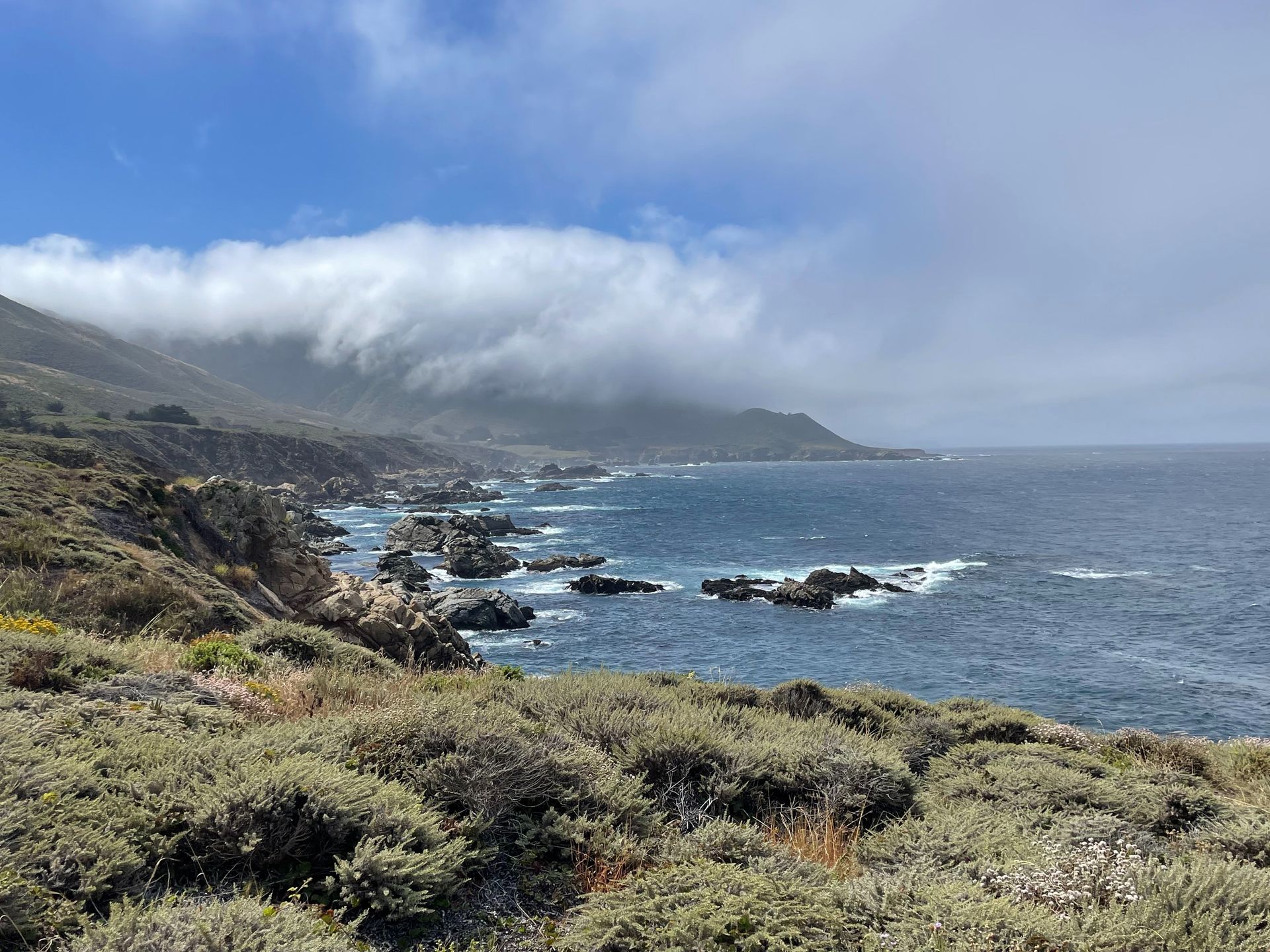 Rocky coastline with ocean waves, cloudy sky, and green hills fading into mist