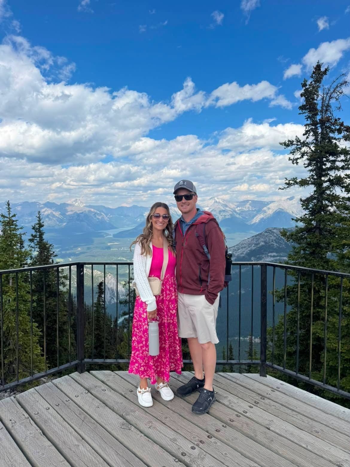 Two people standing on a mountain lookout deck with snowy peaks and blue sky in the background
