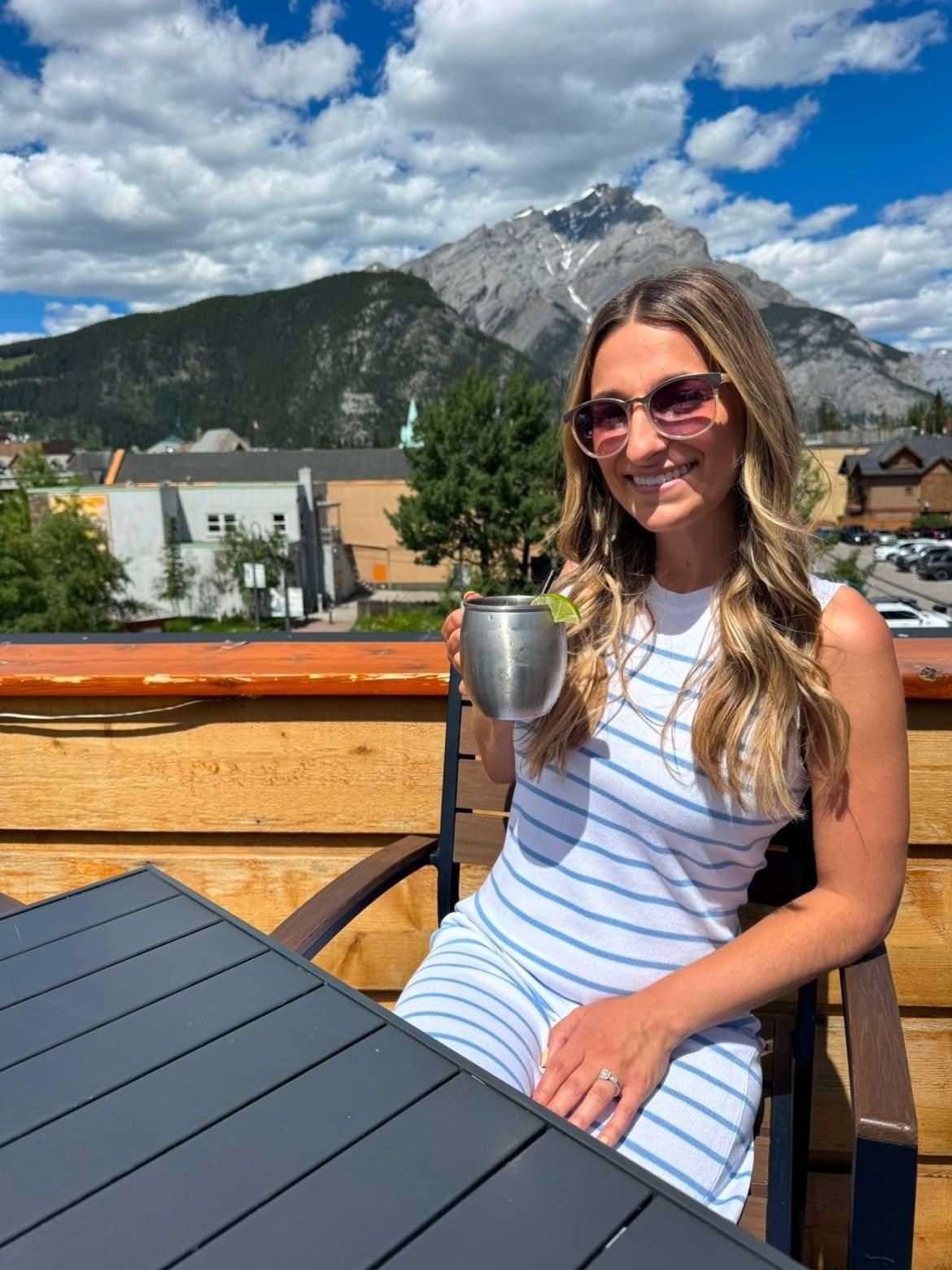 Woman in striped dress and sunglasses holding a cup on a rooftop terrace with mountains in the background