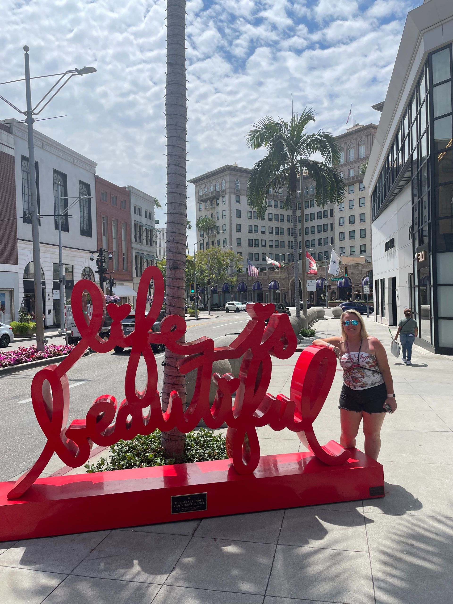 Red “life is beautiful” sculpture on a sunny city sidewalk, with pedestrians and palm trees nearby