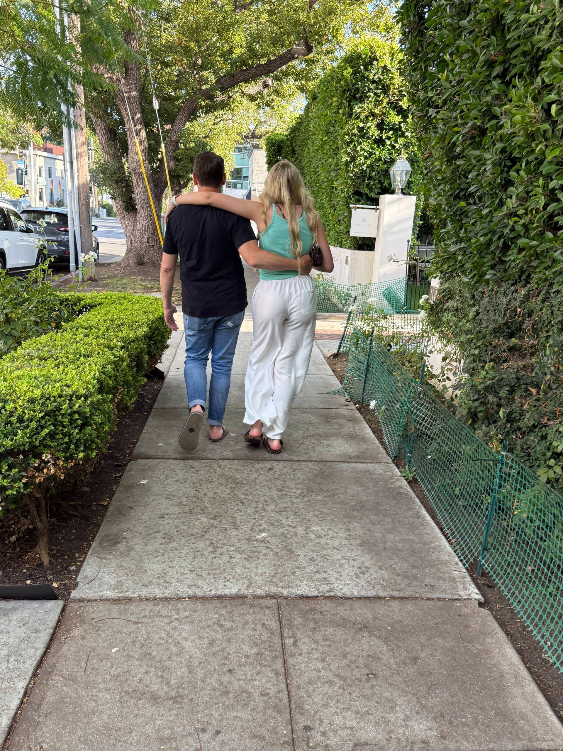 Two people walking arm-in-arm on a sunny sidewalk lined with greenery and hedges.