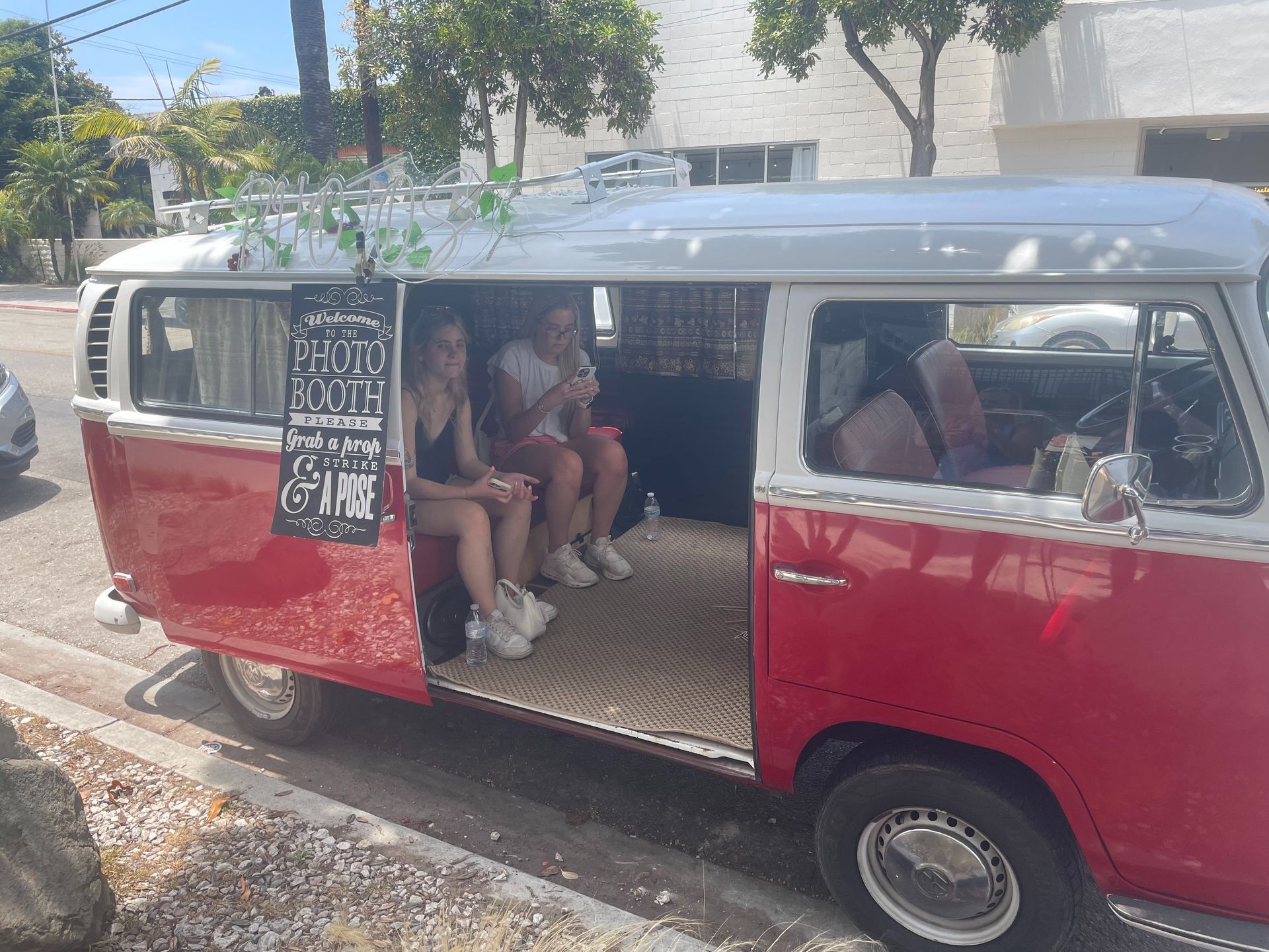 Red vintage van parked curbside with people seated inside and a chalkboard sign on the door.