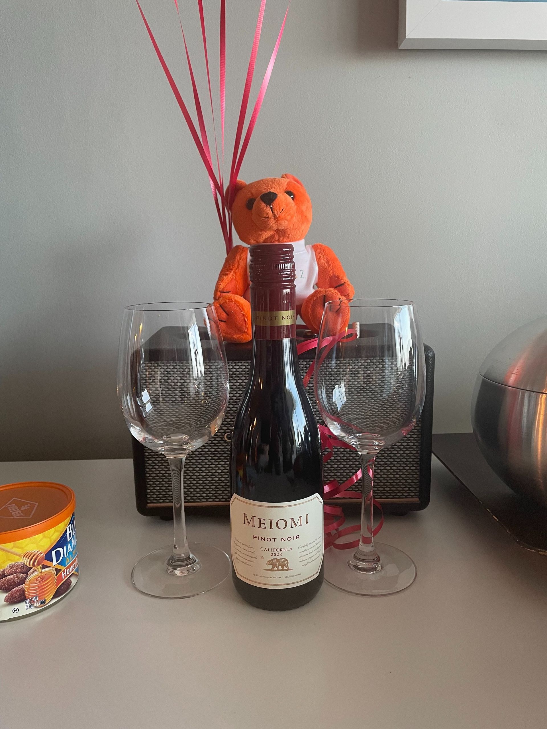 Orange plush toy behind a wine bottle, flanked by two wine glasses on a table with decorative sticks.