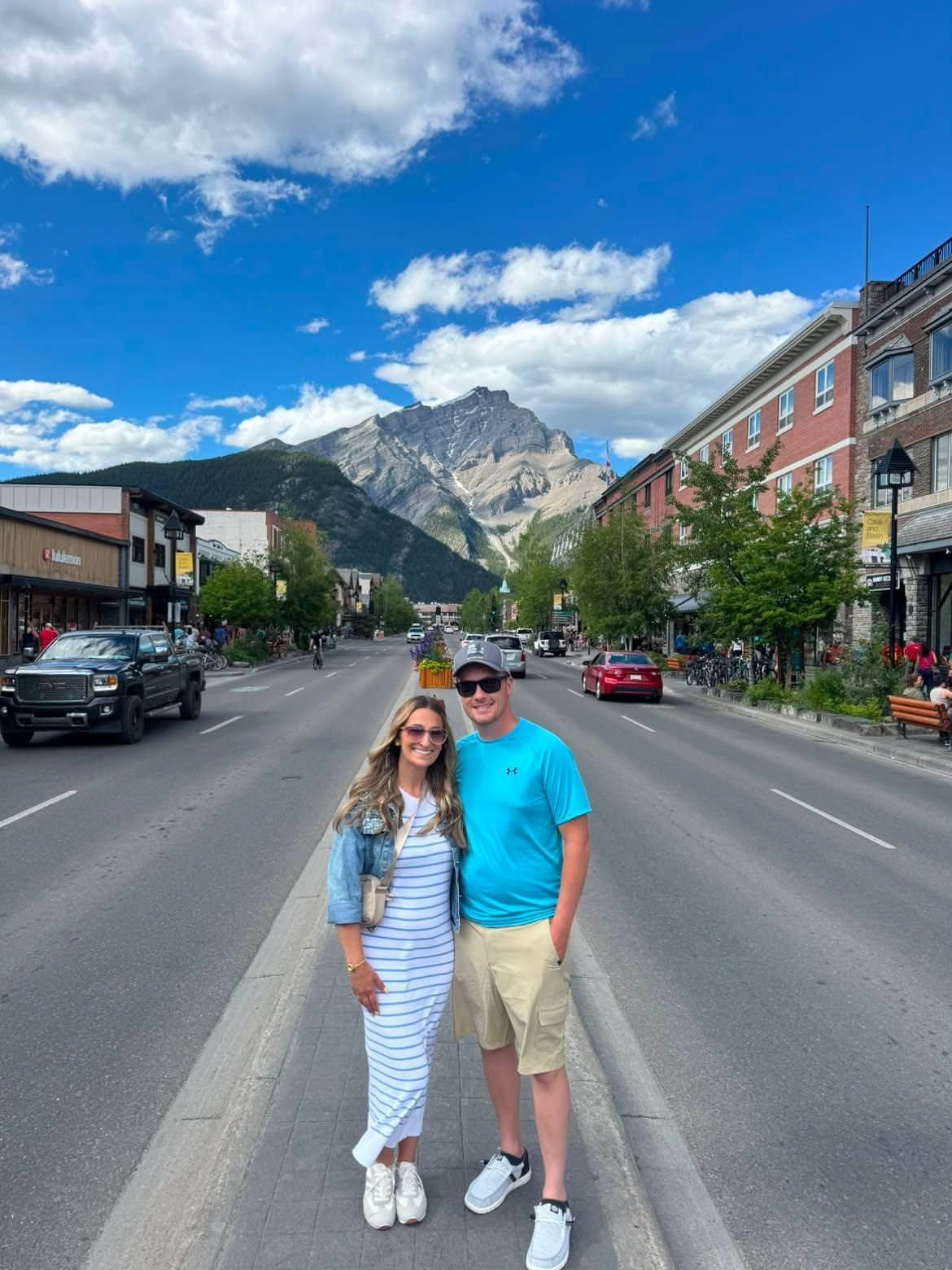 Couple posing on a mountain town street with cars, trees, and rocky peaks under a blue sky.