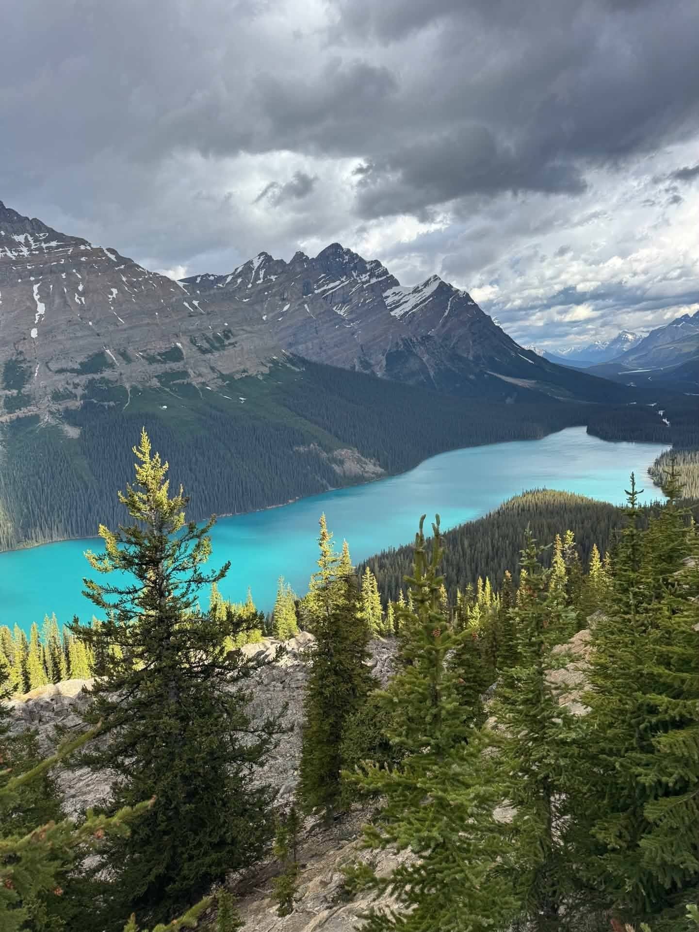 Turquoise alpine lake beneath rugged mountains and cloudy sky, framed by evergreen forest