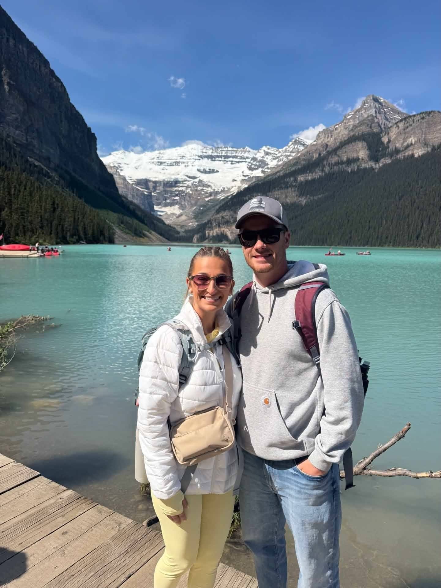 Couple standing by a turquoise alpine lake with mountains and snow-capped peaks in the background