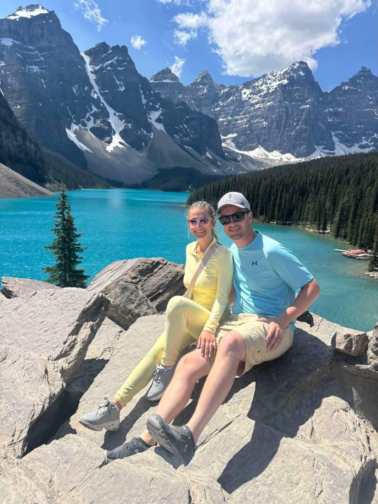 Two people sitting on a rock by a turquoise alpine lake with snowcapped mountains and blue sky in the background