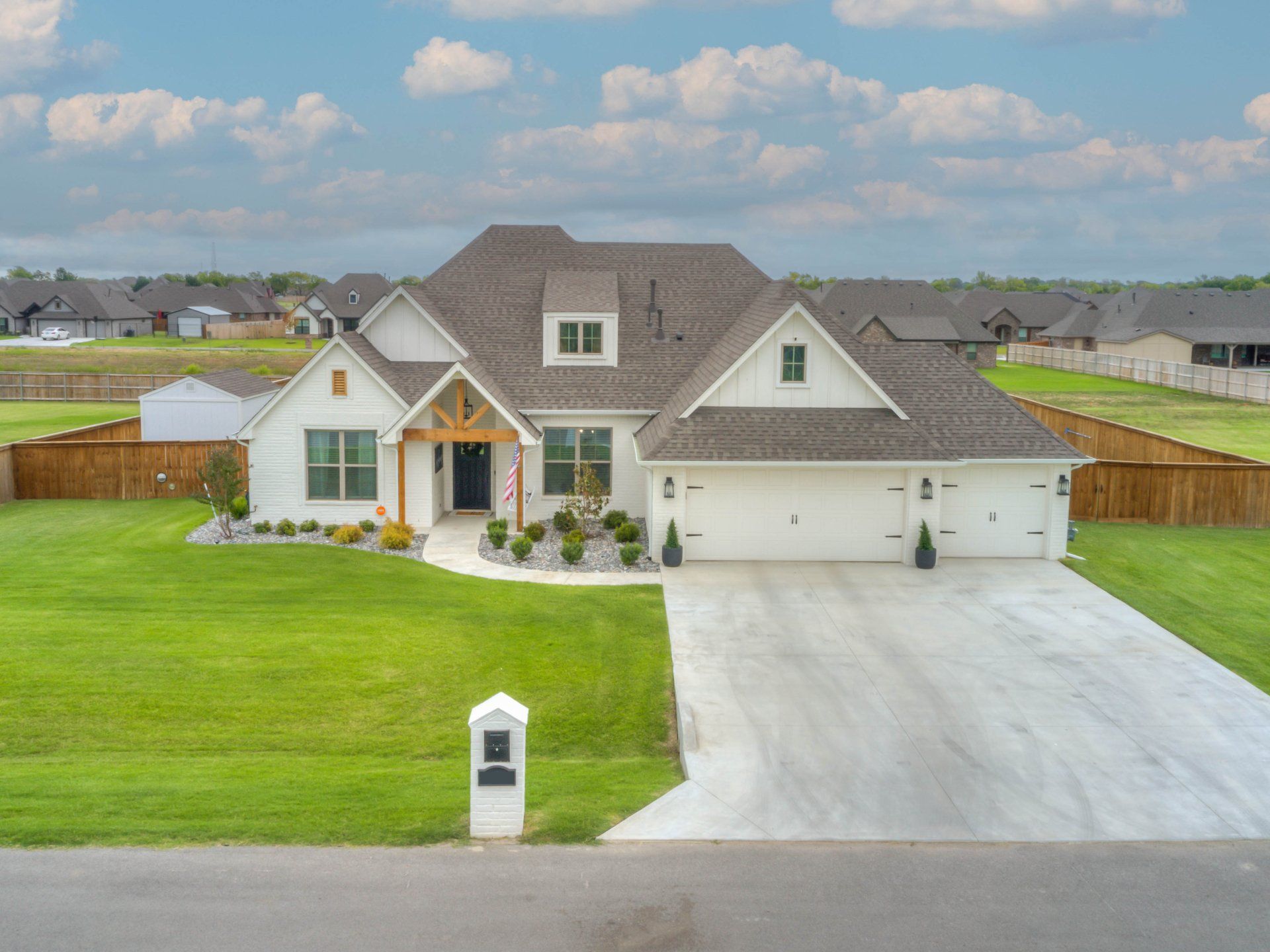 A white, single-story house with brown shingled roofs, a concrete driveway, and a front lawn under a partly cloudy sky.