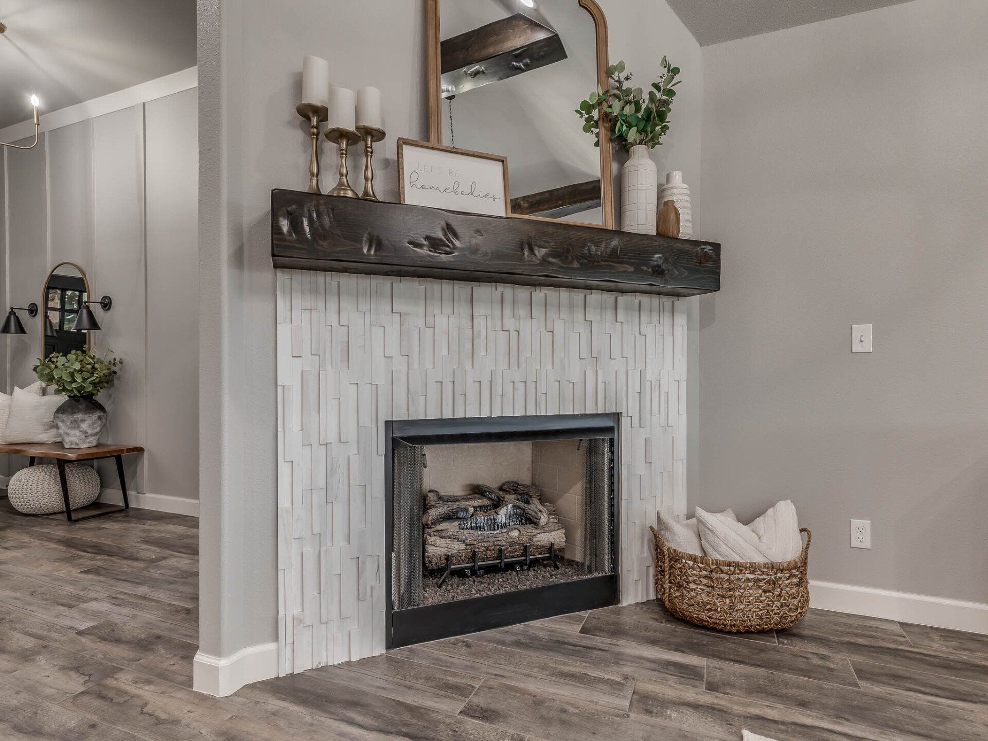 A fireplace with a rustic dark wood mantel, white textured tile surround, mirror, decor, and a woven basket of pillows.