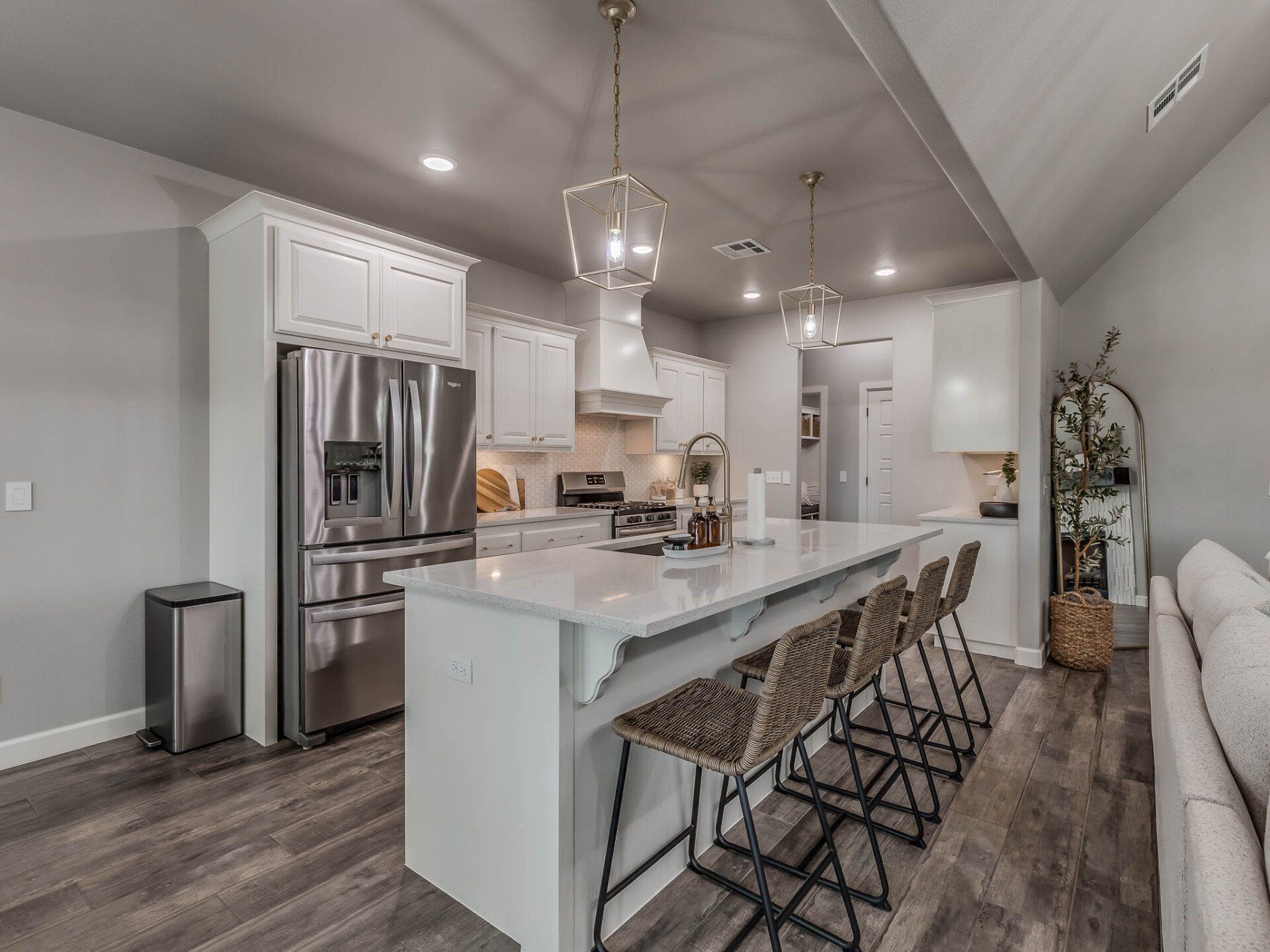 A modern, bright kitchen with white cabinetry, a large marble-topped island, stainless steel fridge, and wood flooring.