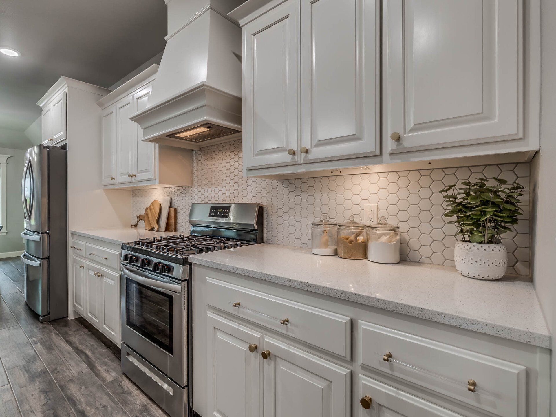 A white kitchen with a stainless steel stove, range hood, and subway tile backsplash, featuring light gray wood floors.
