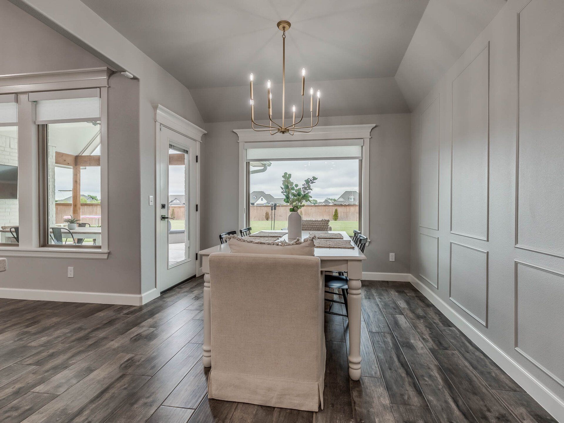 A dining area with a wood table, cream chair, chandelier, and gray wainscoted walls overlooking a backyard.