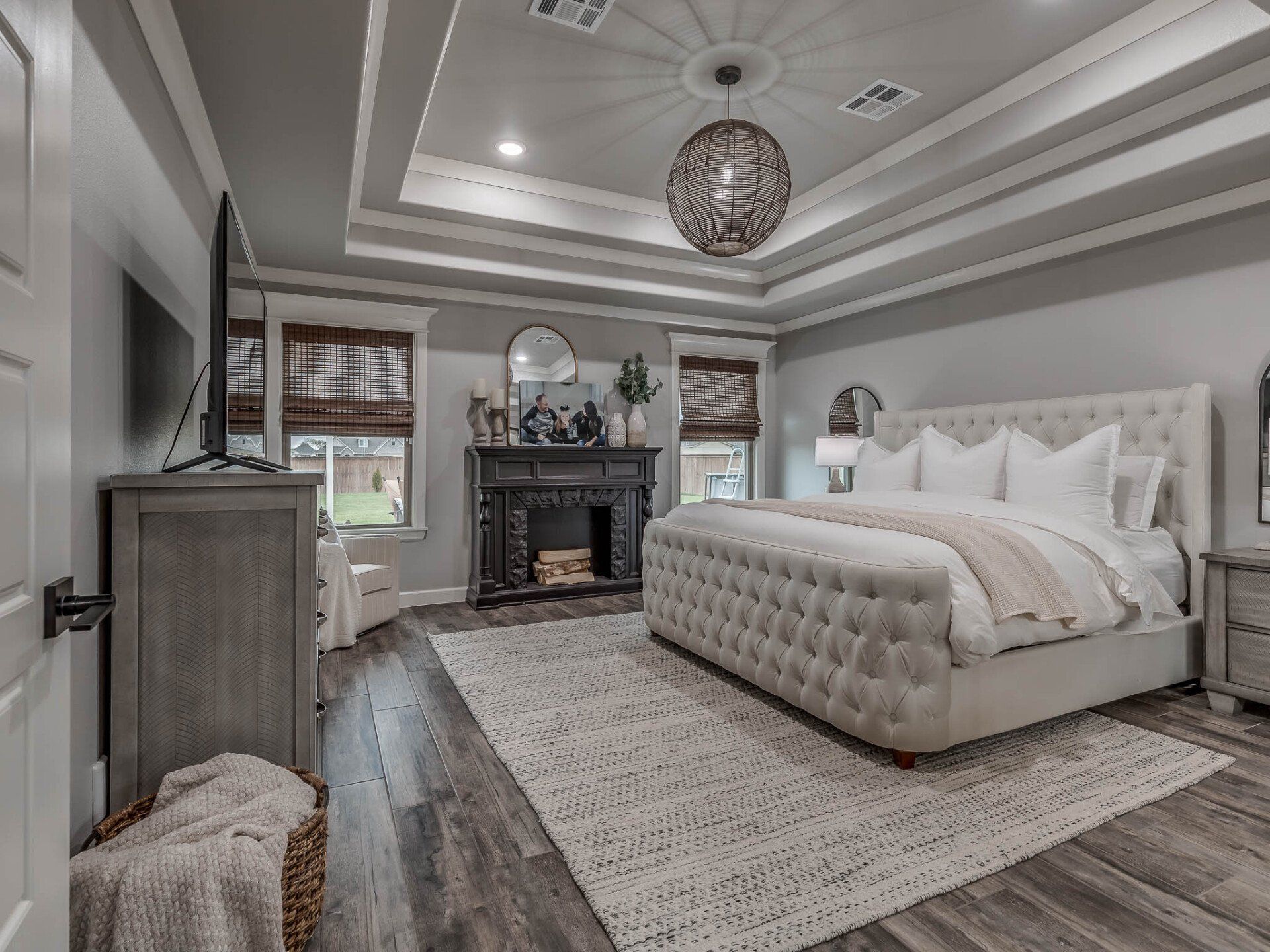 Modern, neutral-toned bedroom with a tufted beige bed, ornate fireplace, tray ceiling, and light gray wood flooring.