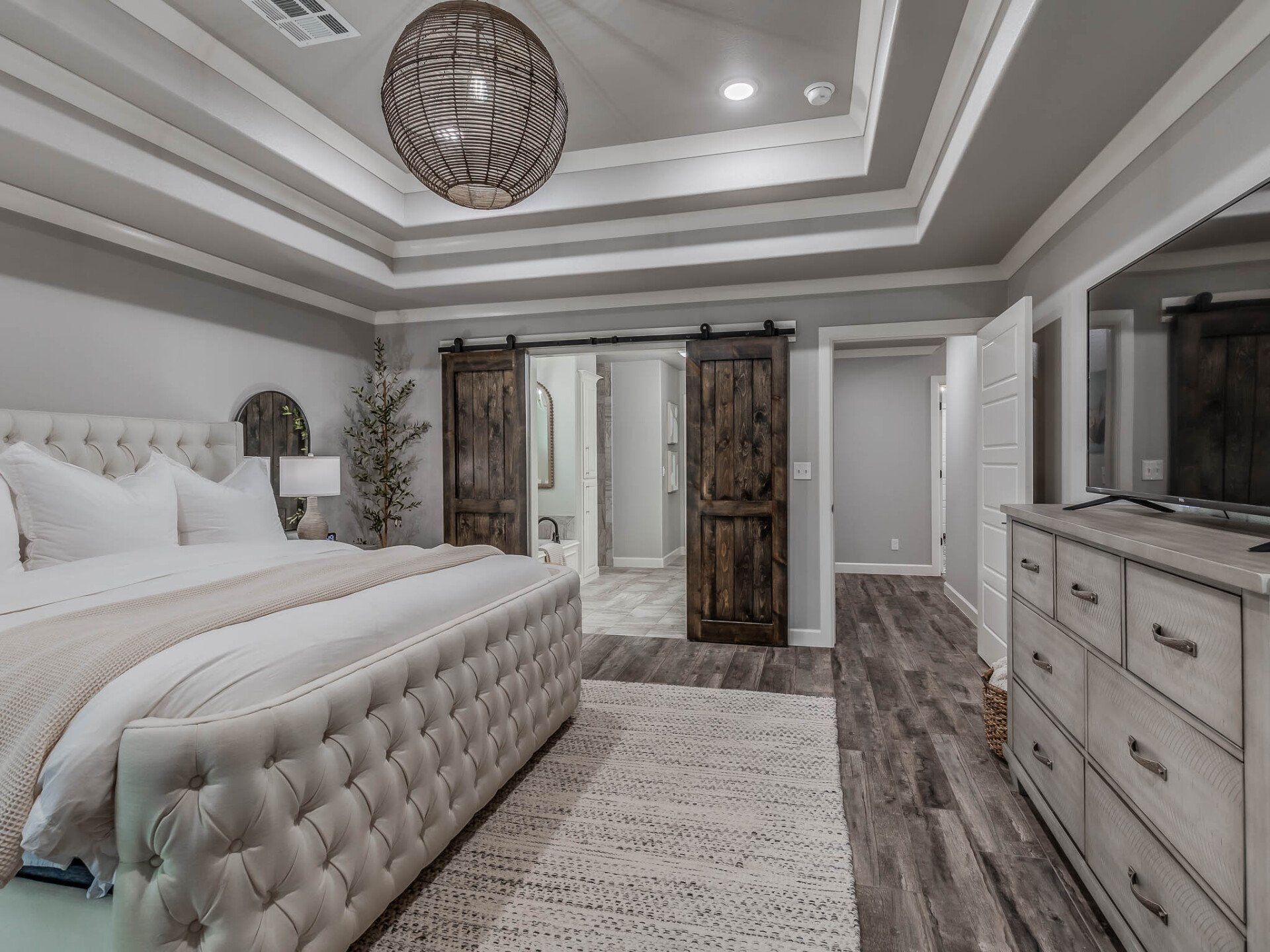 A modern master bedroom features a tufted cream bed, wood sliding barn doors, gray walls, and a large central chandelier.