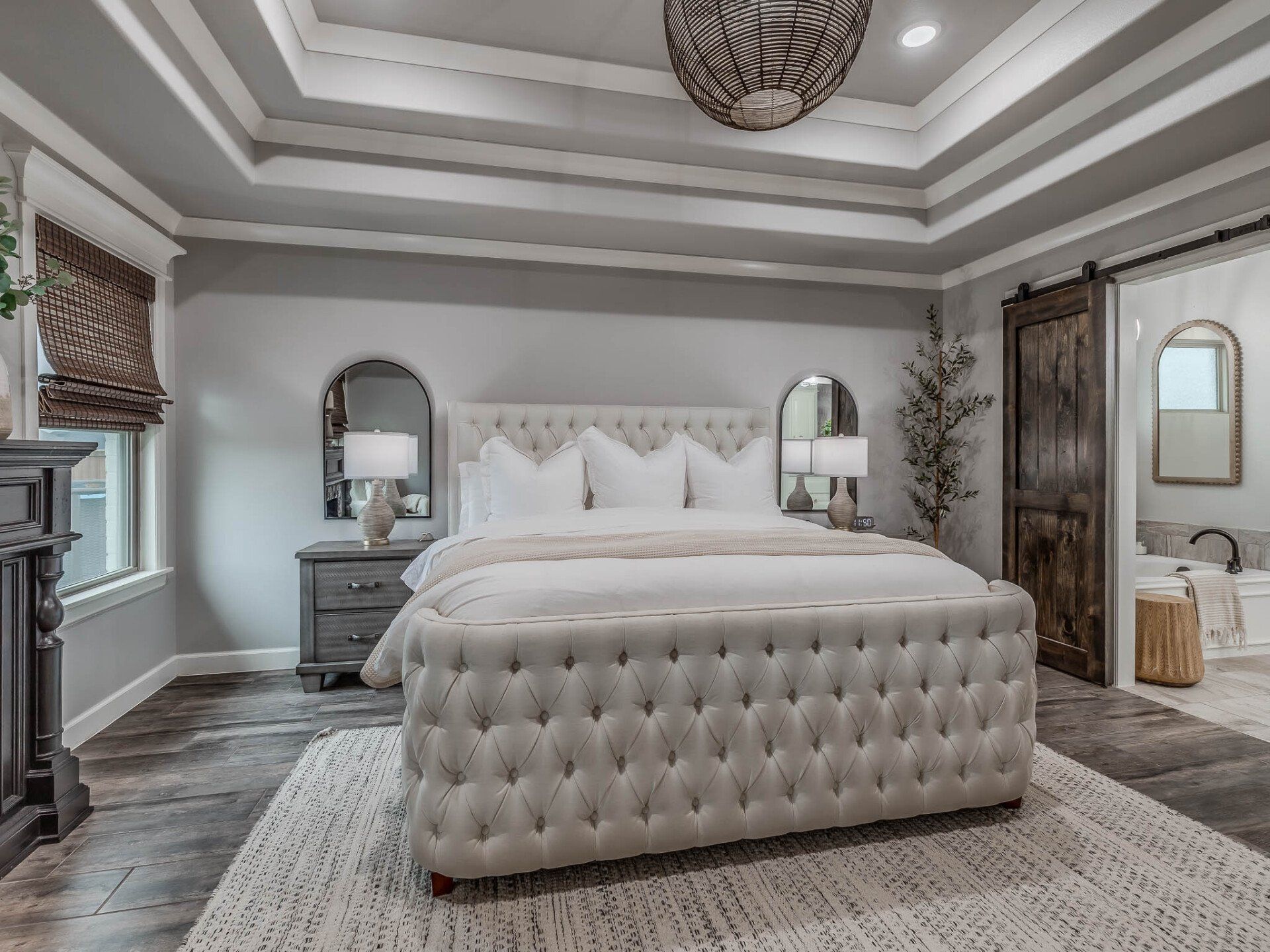 A luxury bedroom with a tufted beige bed, tray ceiling, dark wood floors, and a sliding barn door leading to a bathroom.
