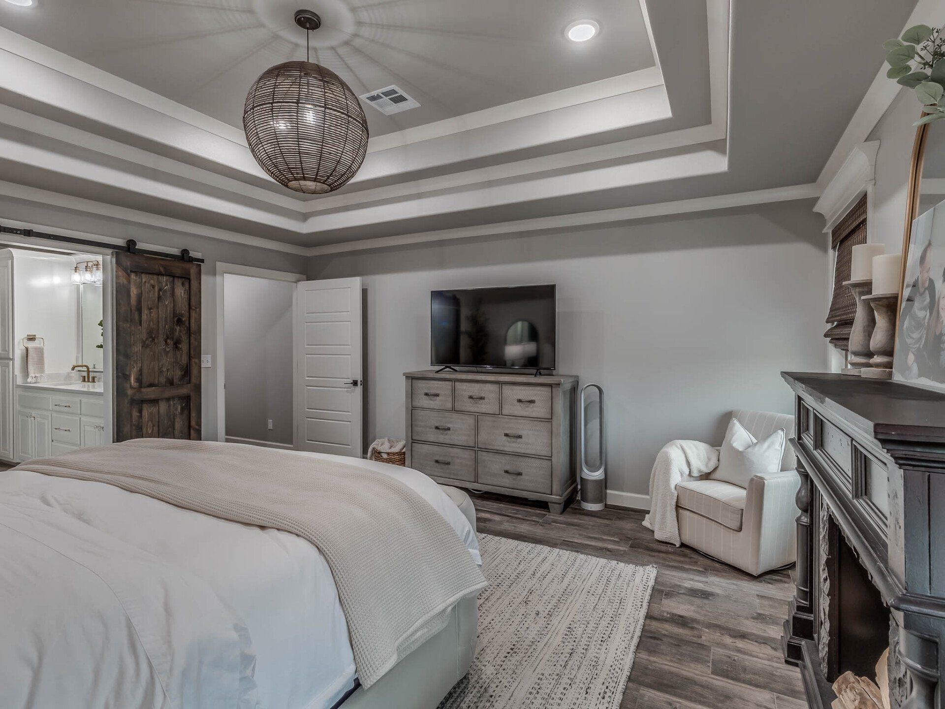 A modern bedroom featuring a tray ceiling, wood-toned furniture, a television on a dresser, and an armchair by a fireplace.