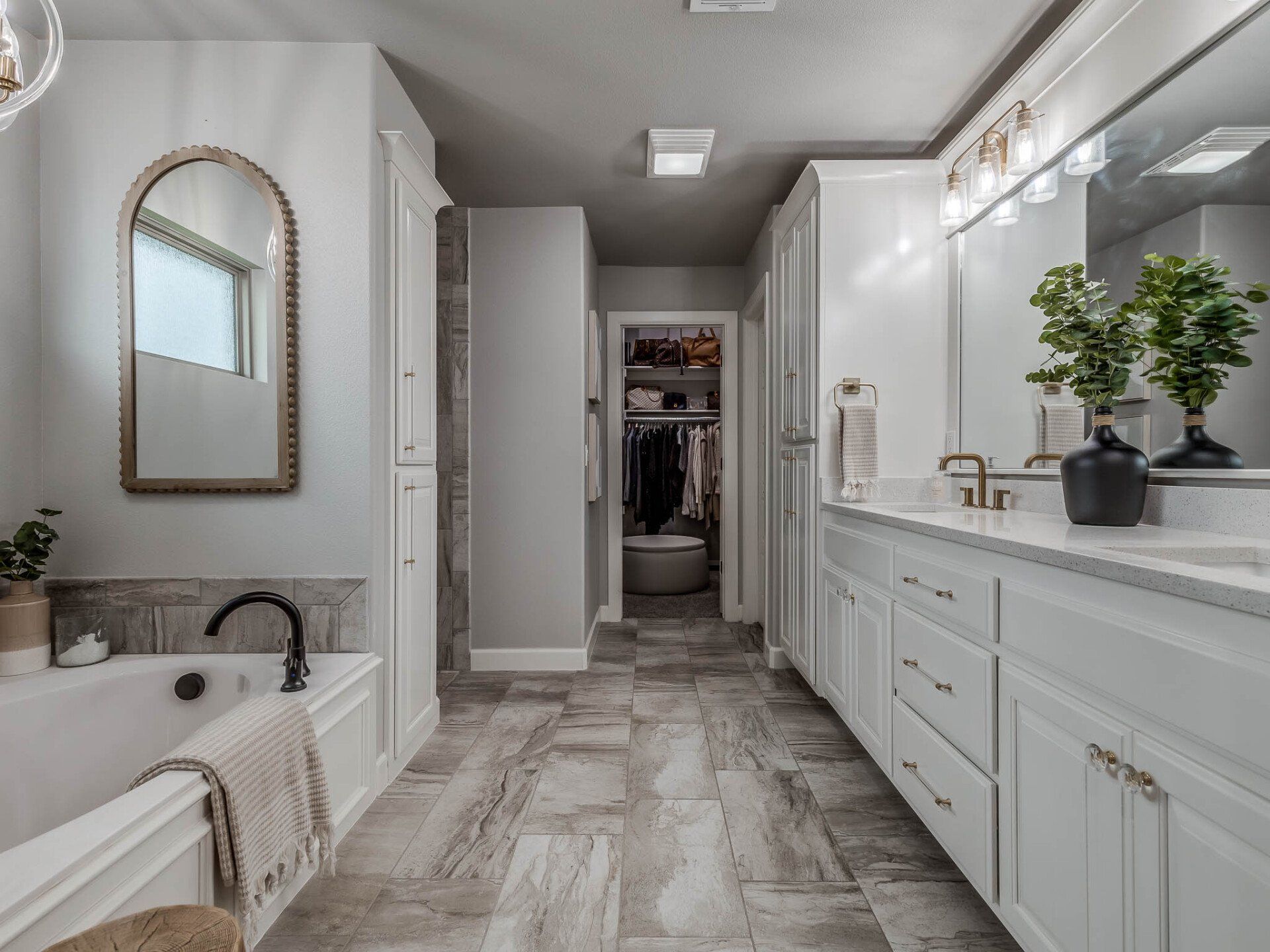 A bright, modern bathroom with a white soaking tub, double vanity, grey stone-look tile flooring, and a walk-in closet.