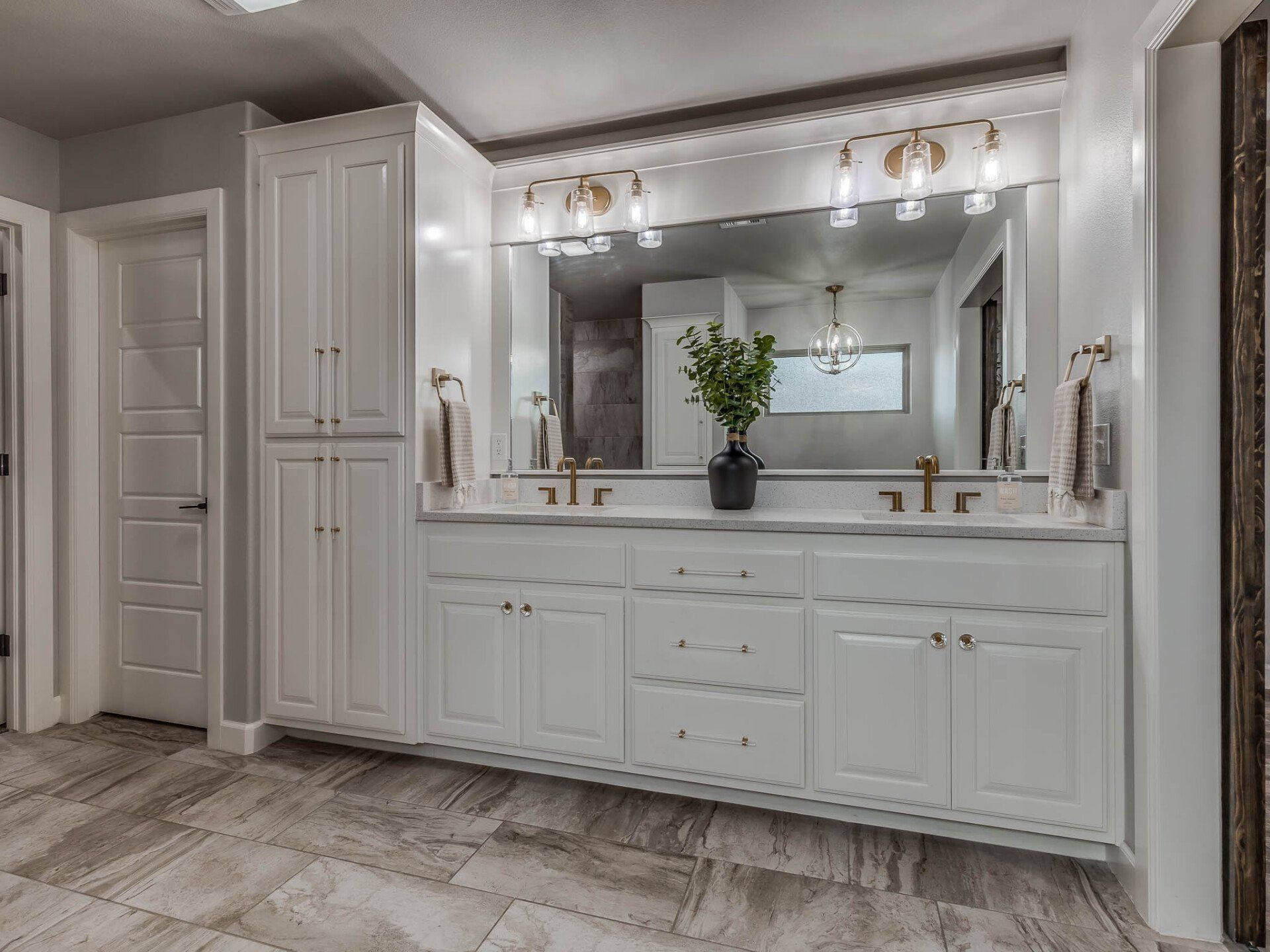 A bright white double-vanity bathroom with gold hardware, a large mirror, and tiled flooring.