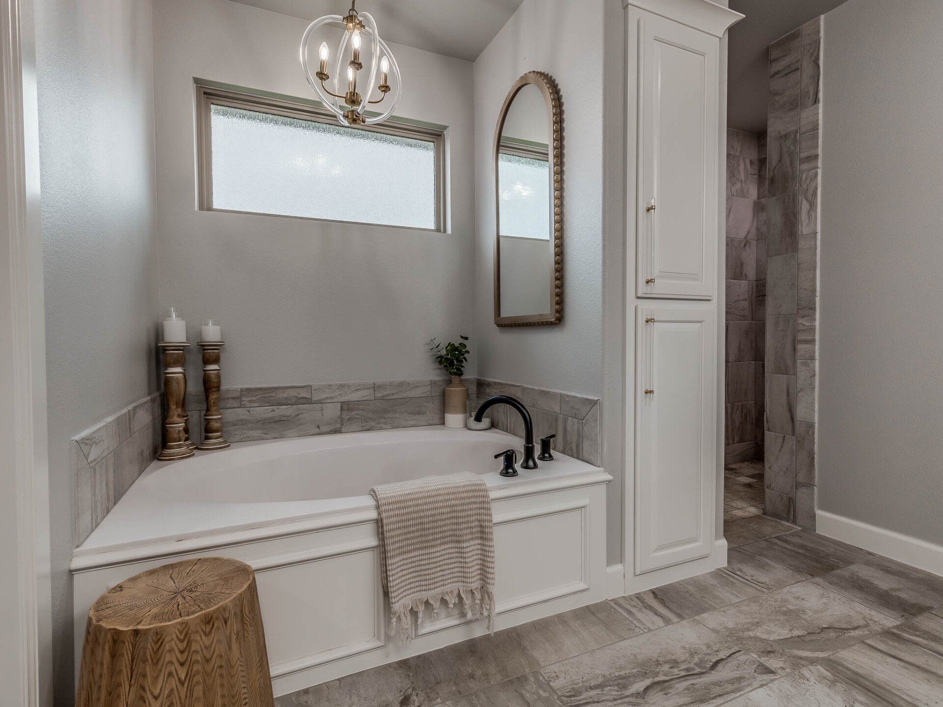 A bright bathroom featuring a white soaking tub, light gray walls, wooden accents, and a tall white cabinet.