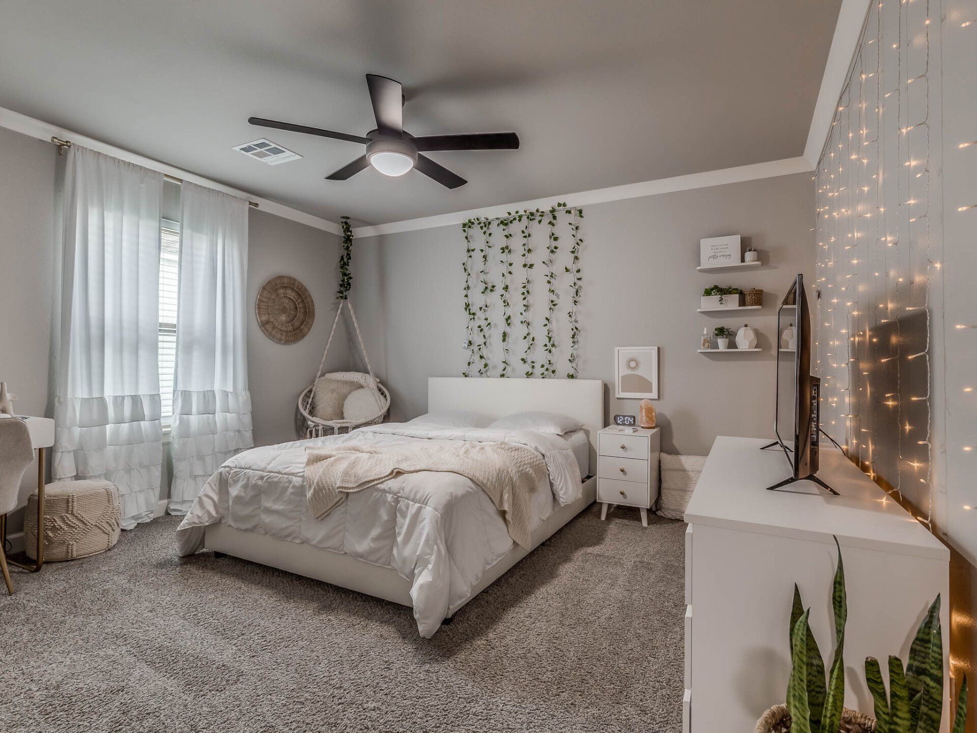 A cozy, neutrally-decorated bedroom featuring a white bed, hanging chair, white dresser, and fairy lights on the wall.
