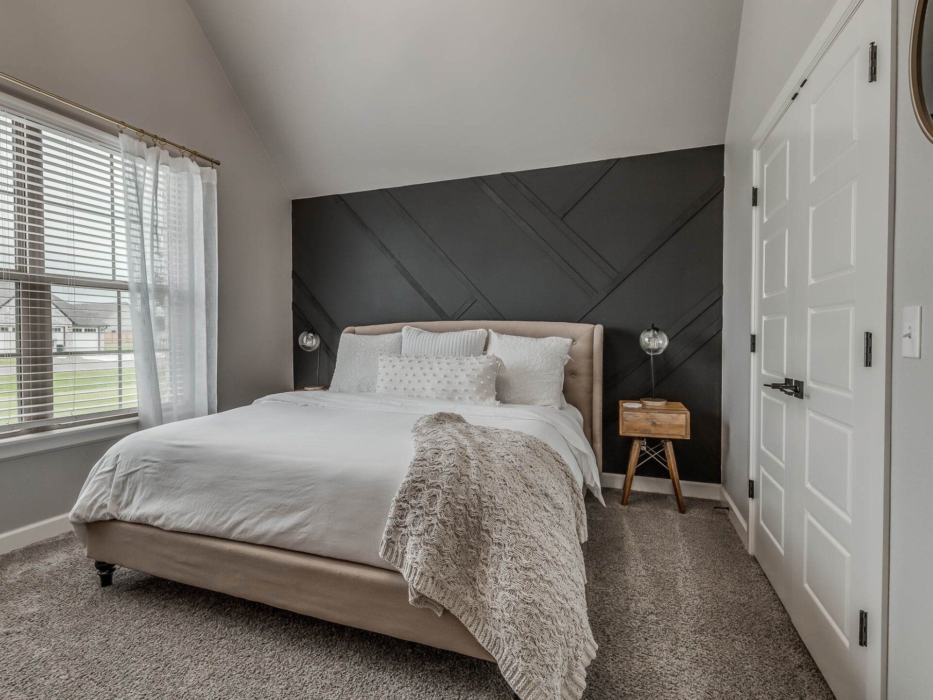 A bedroom with a beige upholstered bed, white bedding, and a black accent wall featuring a geometric wood pattern.