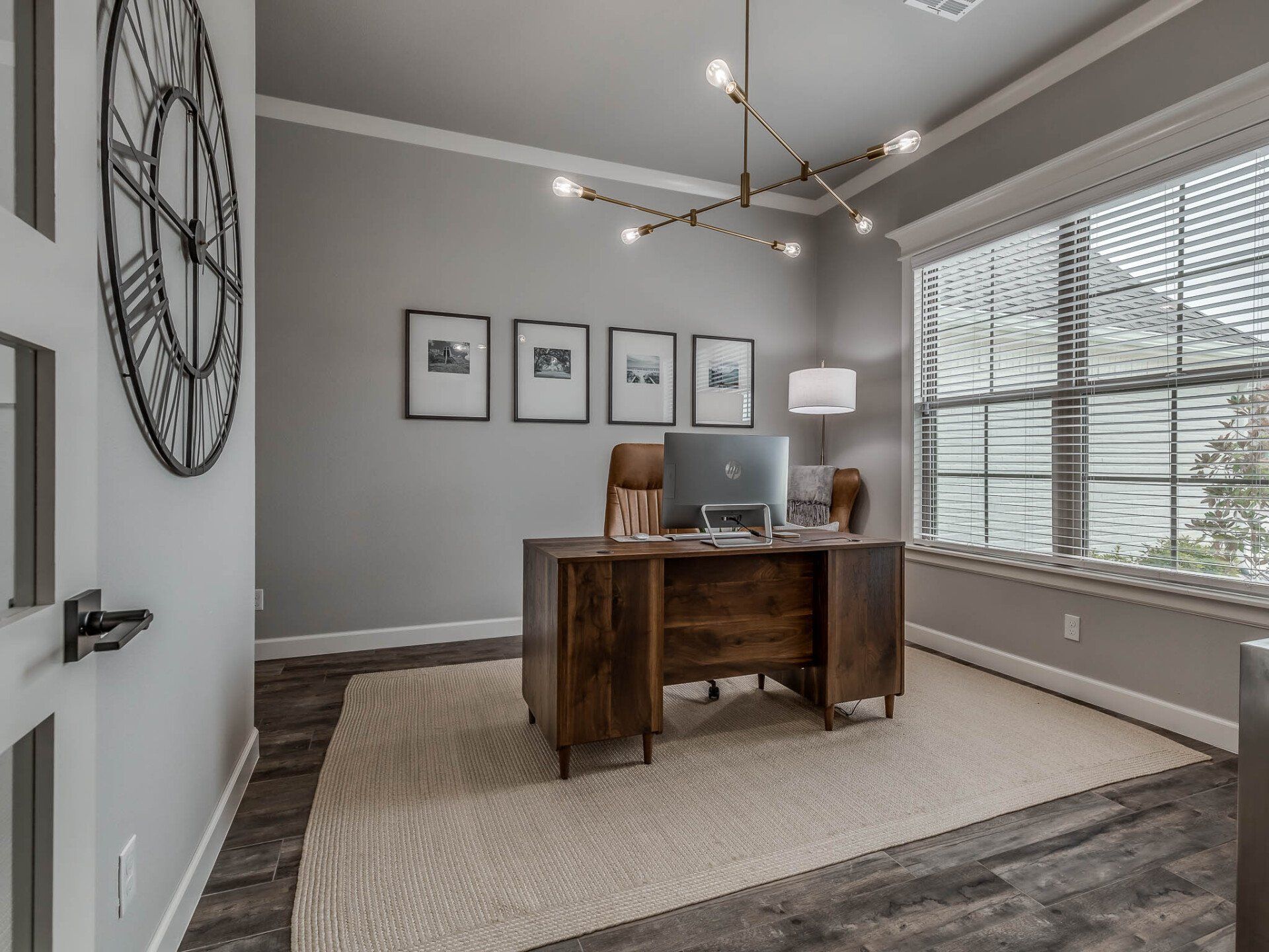 A home office with a dark wood desk, central computer, four framed wall prints, a large wall clock, and a light rug.