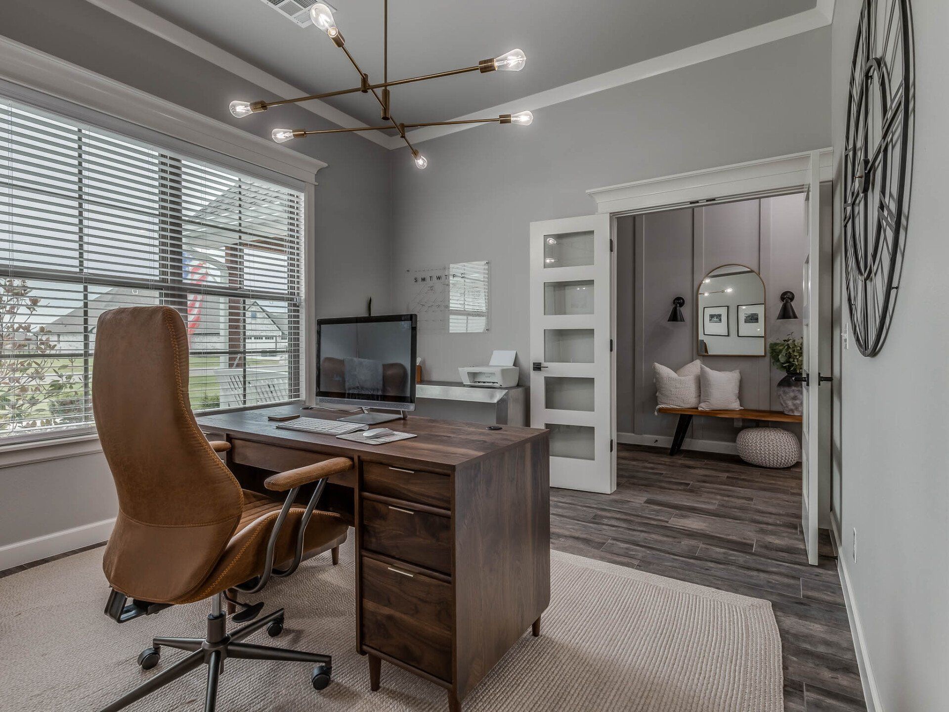 A home office with a brown leather chair at a wooden desk, light gray walls, and an open doorway to a cozy sitting area.