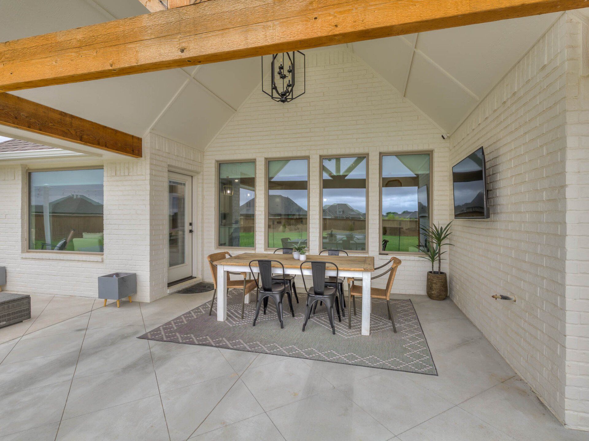 A covered white brick patio featuring a wooden dining table with four chairs on a patterned rug, facing a flat-screen TV.