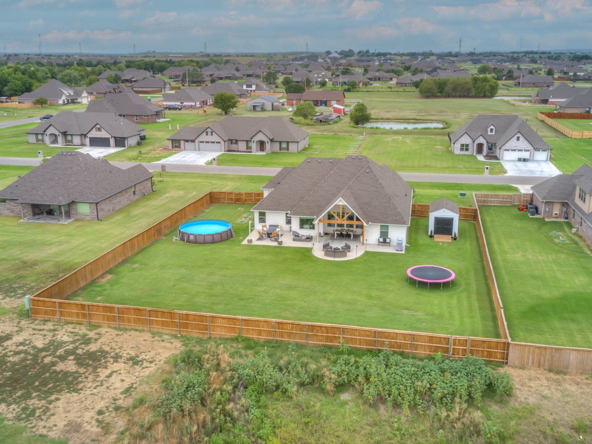 An aerial view of a residential suburban home with a fenced backyard containing a round pool and a trampoline.