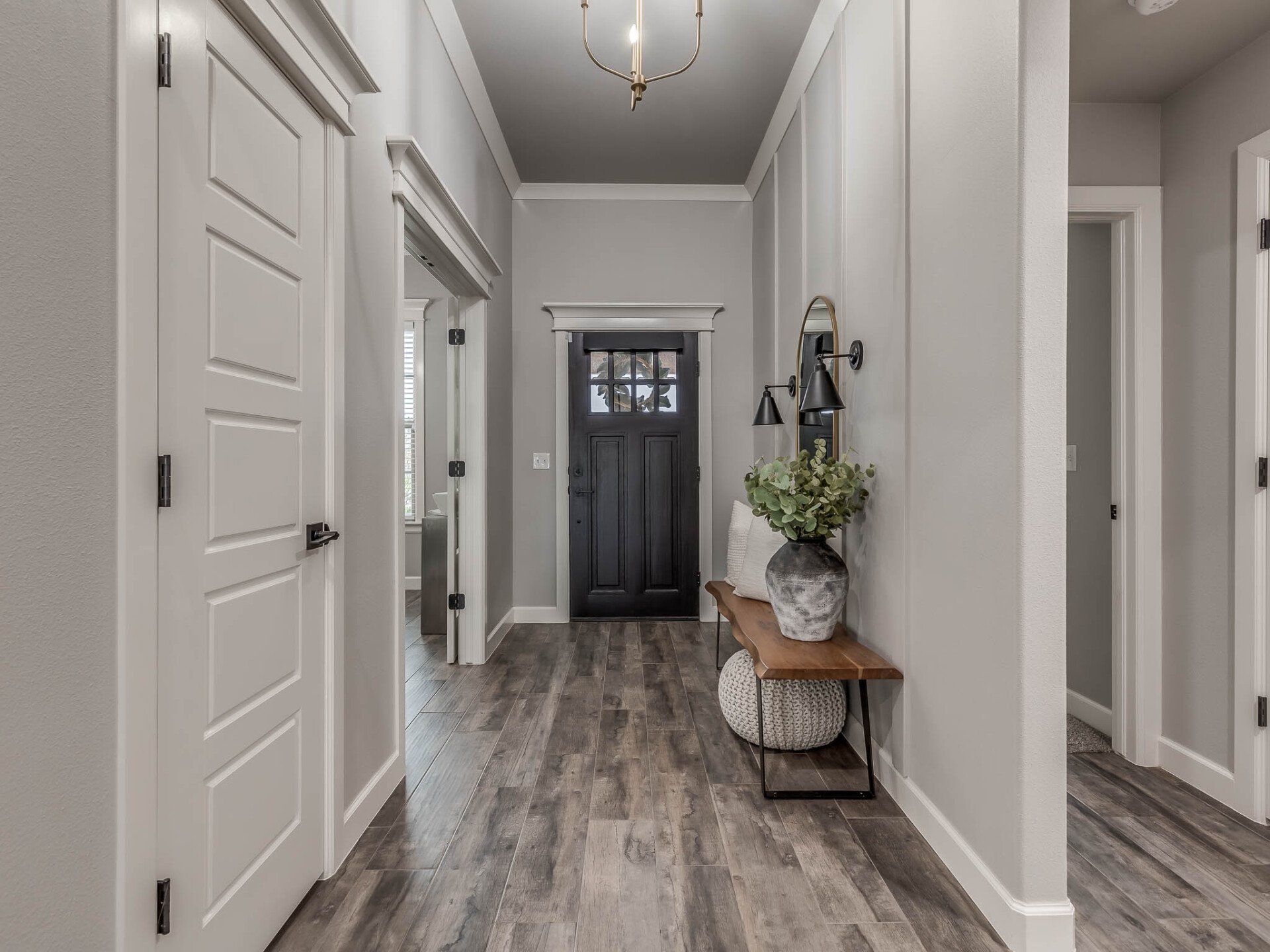 A hallway with light gray walls, wood-look flooring, a black front door, a pendant light, and a wooden bench with decor.