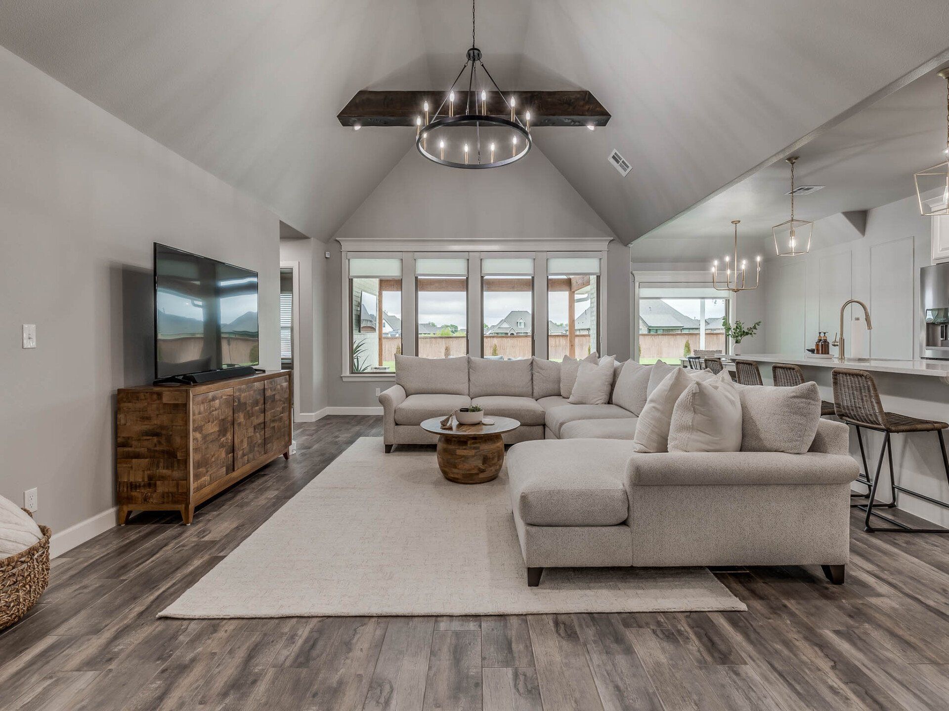 A bright, modern open-plan living room with a large gray sectional, wooden media console, and a vaulted ceiling.