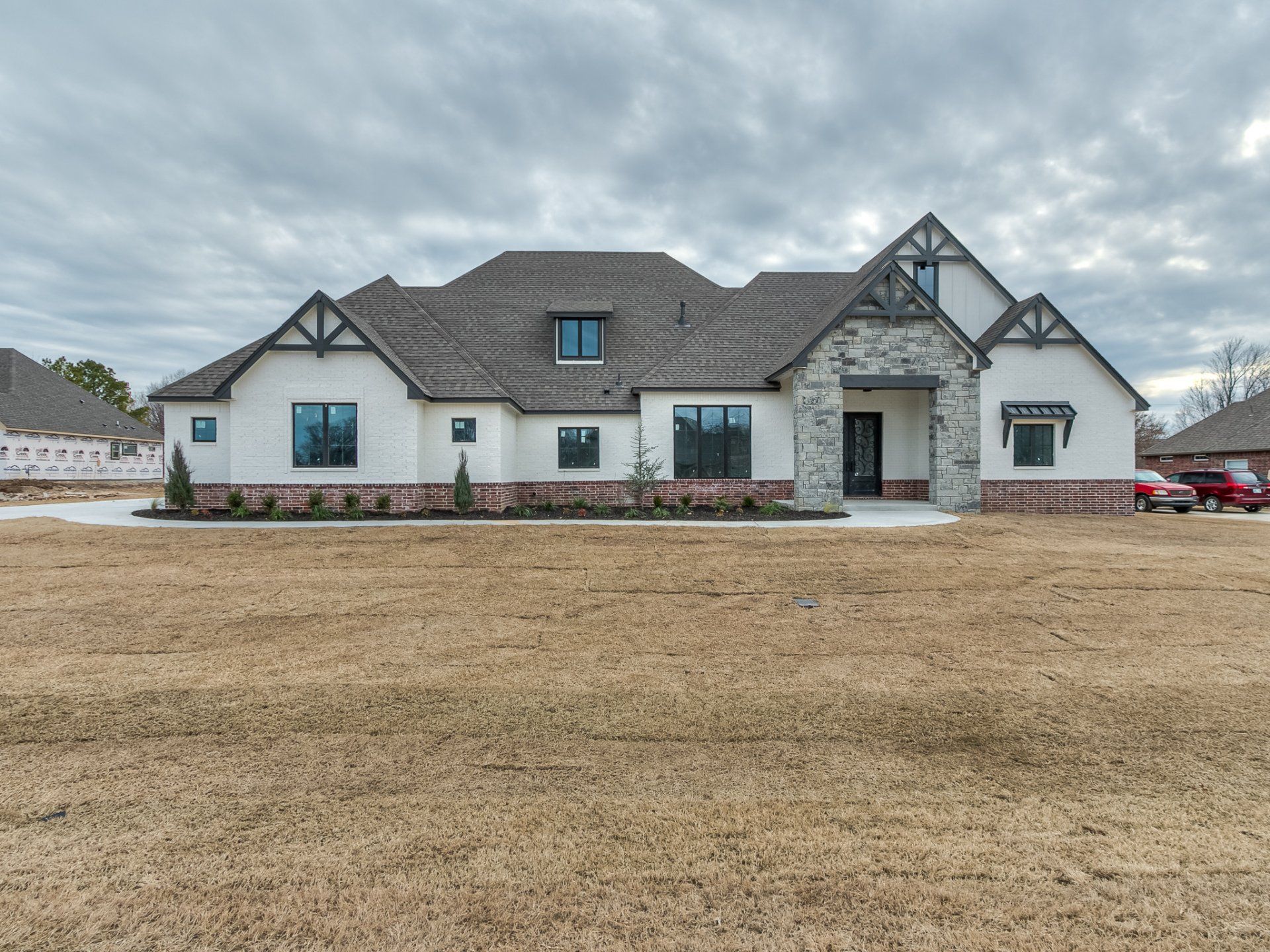 A large, single-story modern home with white stucco, stone accents, brown roof, and a brown lawn under a cloudy sky.